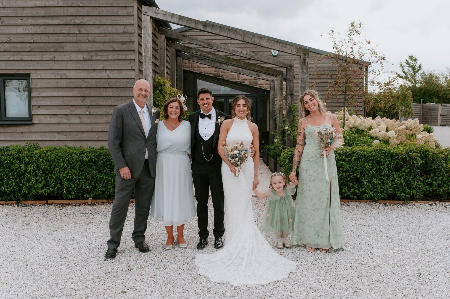 Wedding party members in sage green dresses and formal attire pose together outdoors at rustic venue.