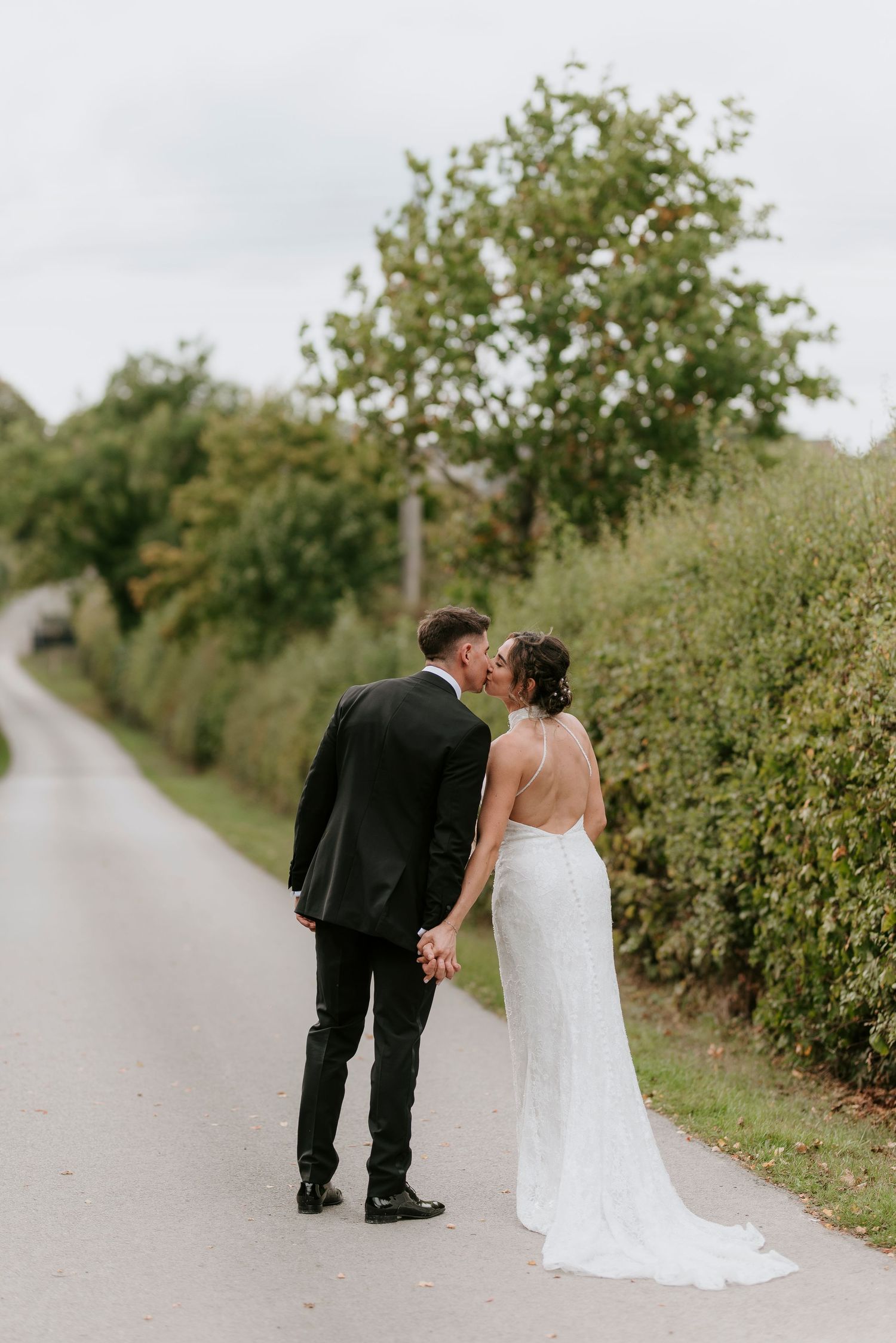 A couple in wedding attire share an intimate moment on a countryside path lined with green bushes.