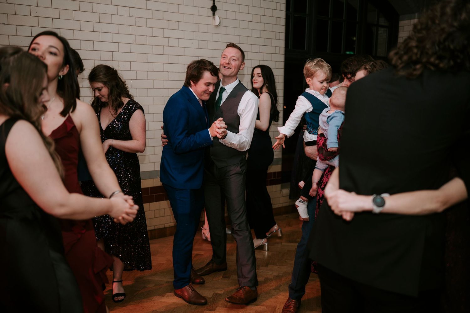 Wedding guests dancing and celebrating on a wooden dance floor at an indoor reception.
