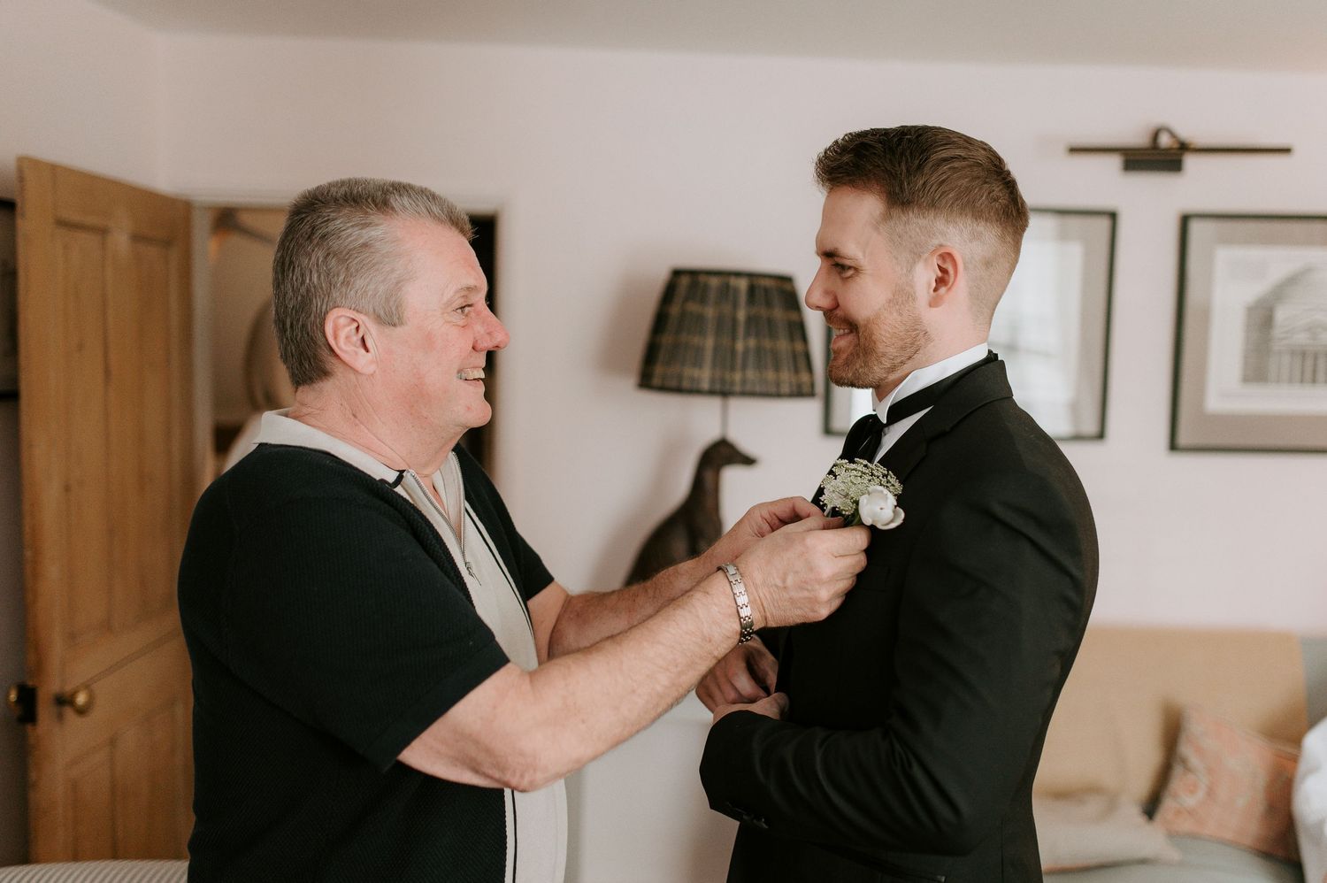 Two people in formal attire help each other with boutonniere pins during a wedding preparation.