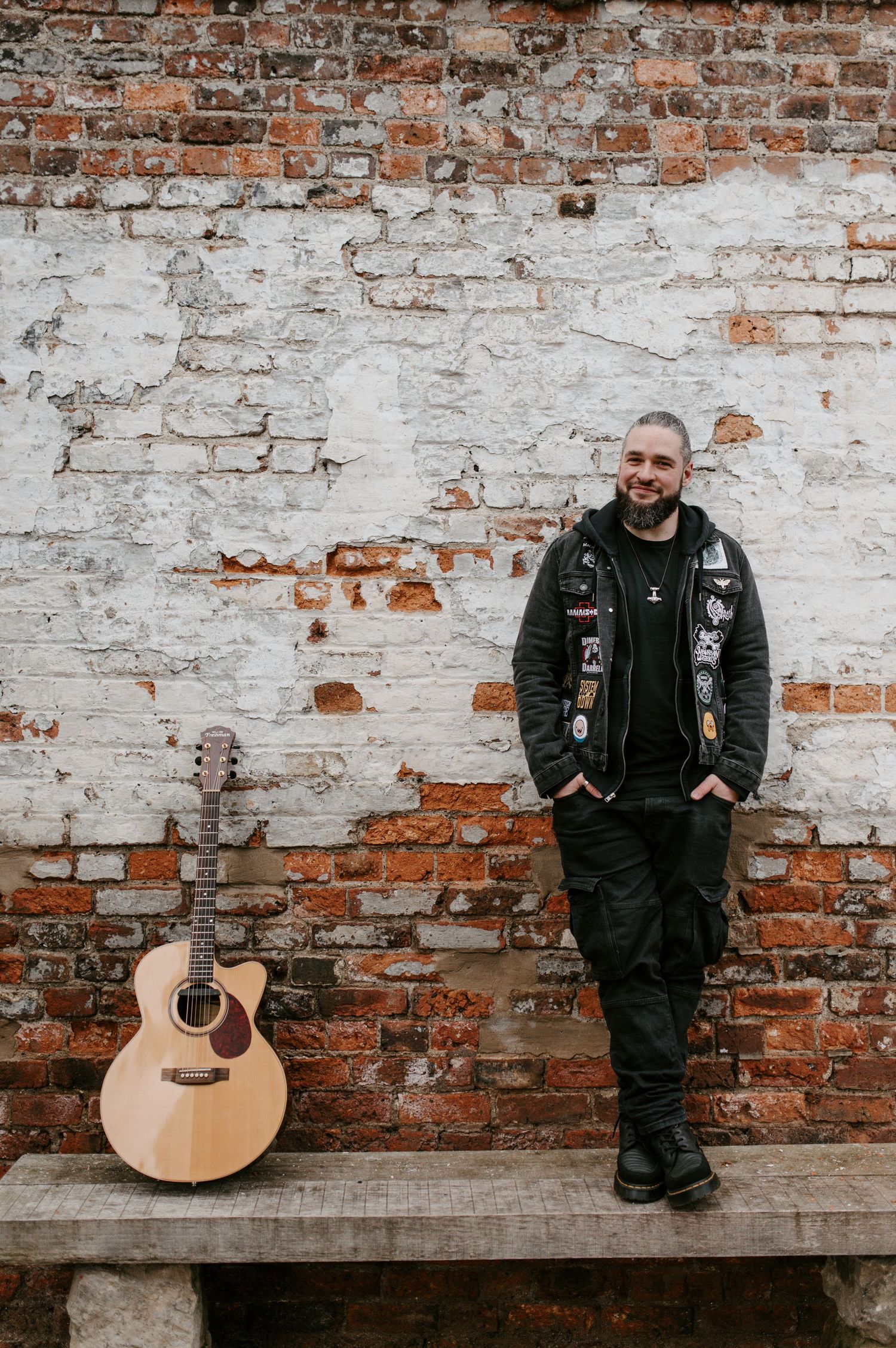 Musician in black leather jacket poses against weathered brick wall with acoustic guitar.