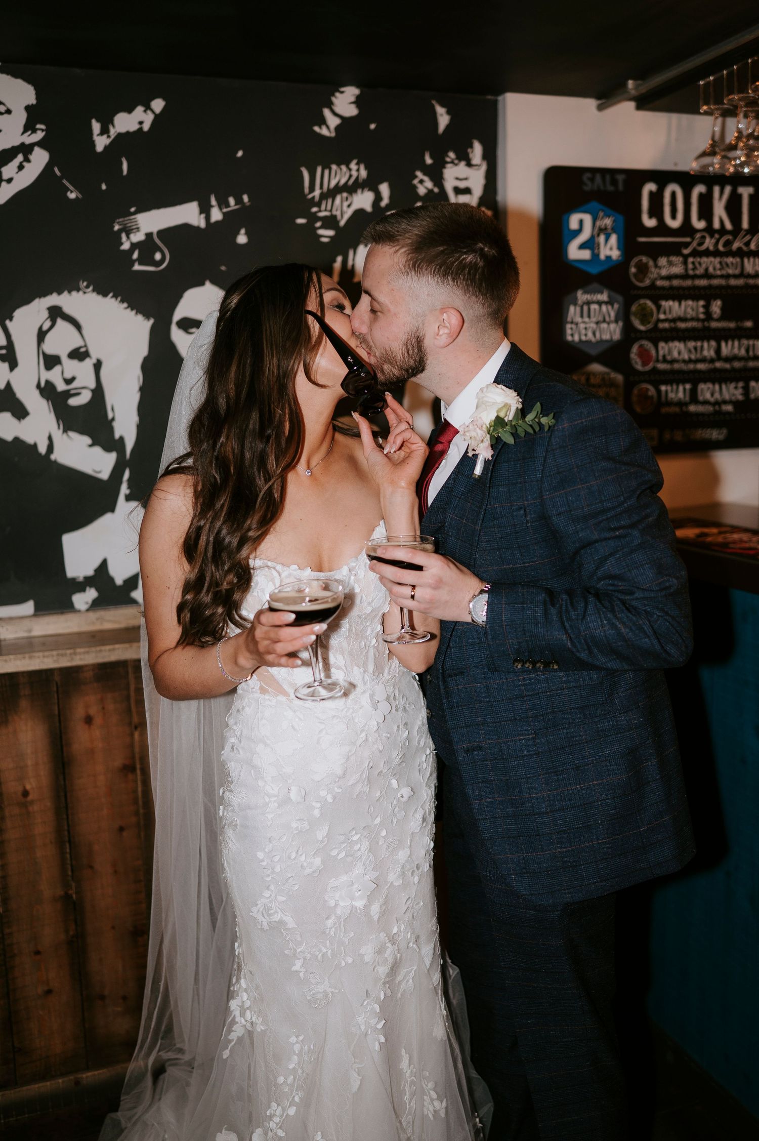 Bride and groom sharing a romantic kiss while holding wine glasses at their wedding reception bar.