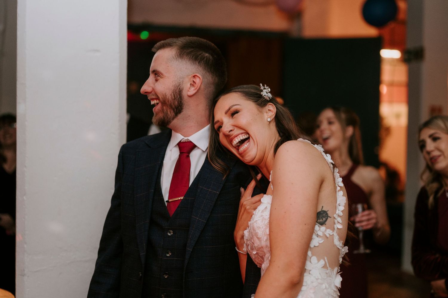Bride and groom laugh together at wedding reception, groom wearing navy suit with red tie.