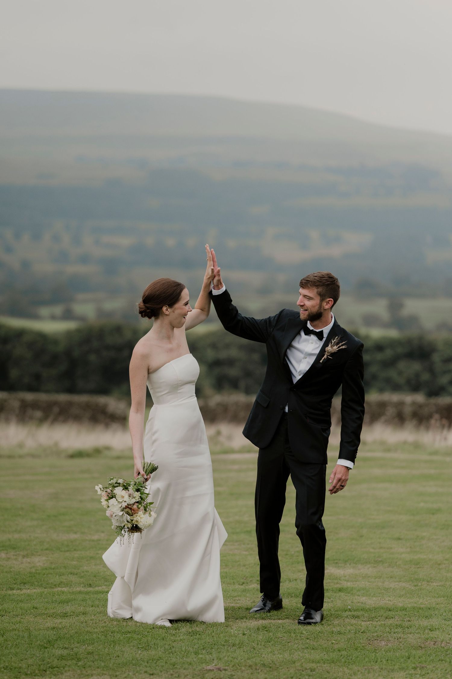 A couple celebrates with a high five on their wedding day against a scenic countryside backdrop.