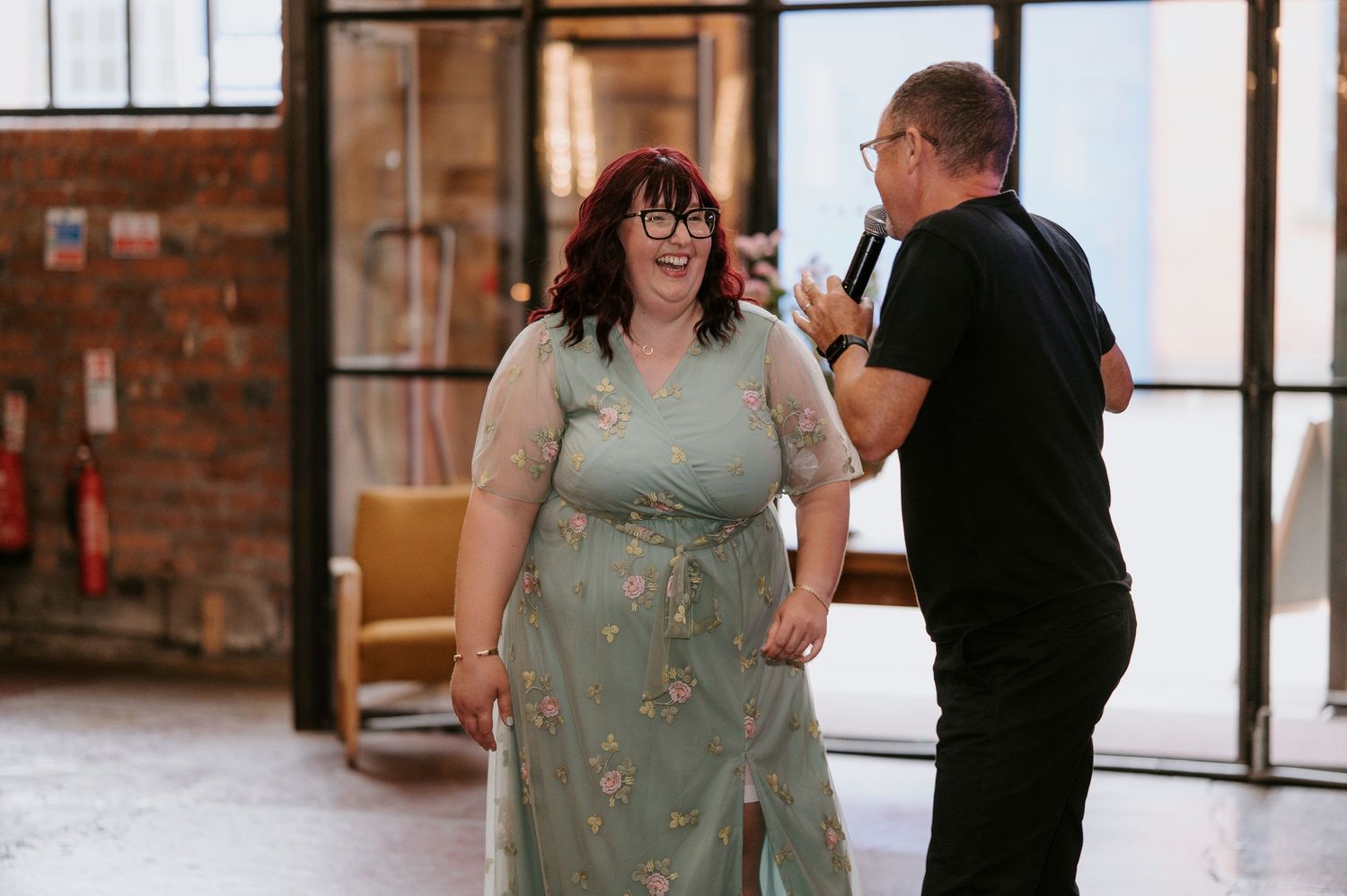 A couple shares a joyful moment dancing together in a sunlit room with brick walls and large windows.