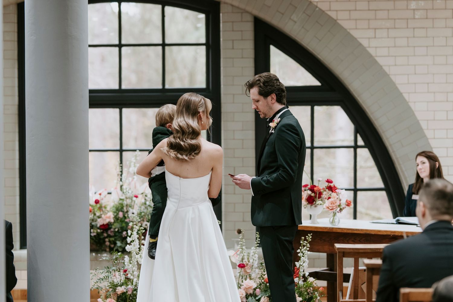 A couple exchanges vows during an indoor wedding ceremony beneath a white brick archway with floral arrangements.