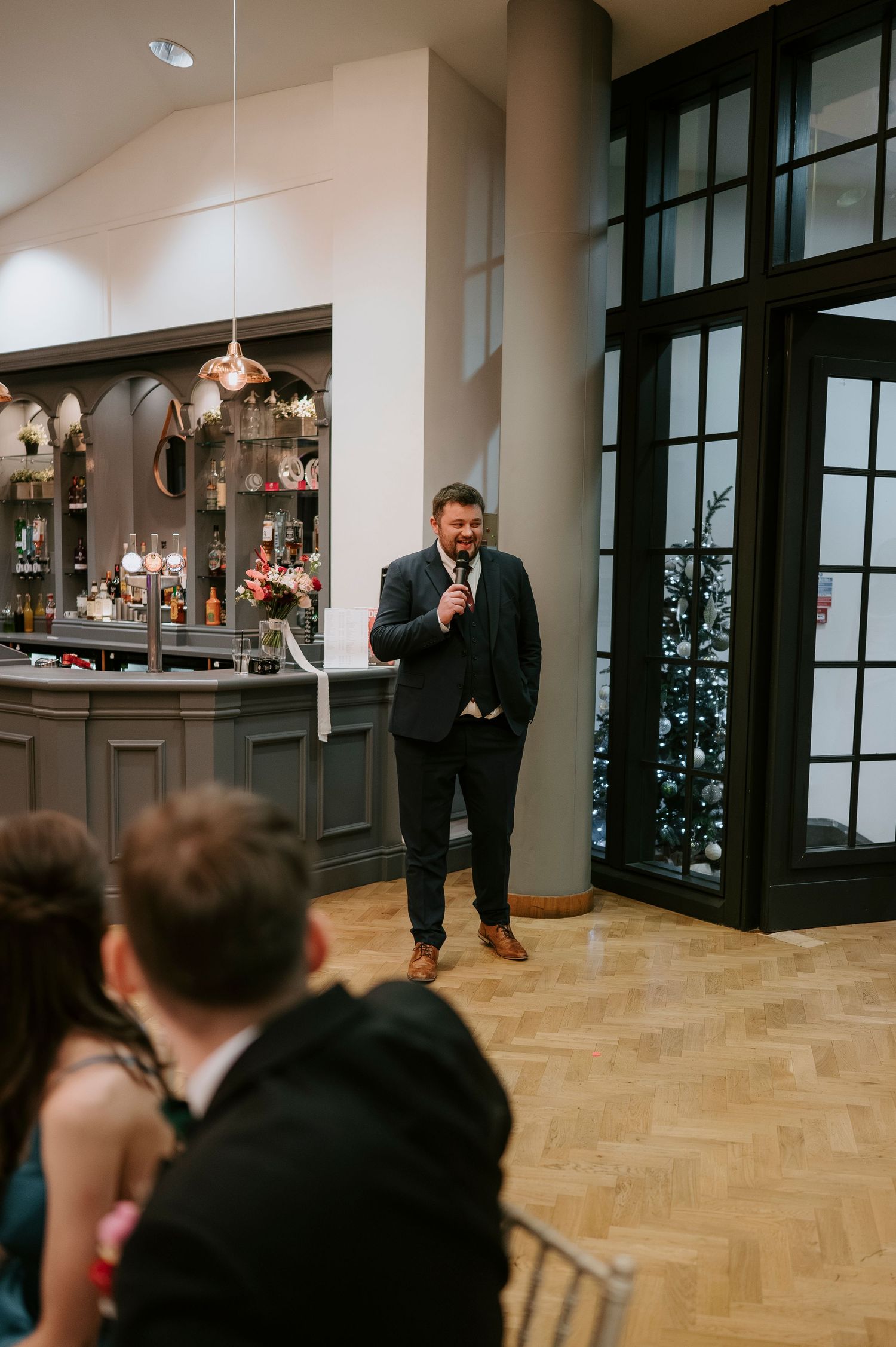 Wedding guests enjoy a speech in an elegant venue with herringbone wood floors and large windows.