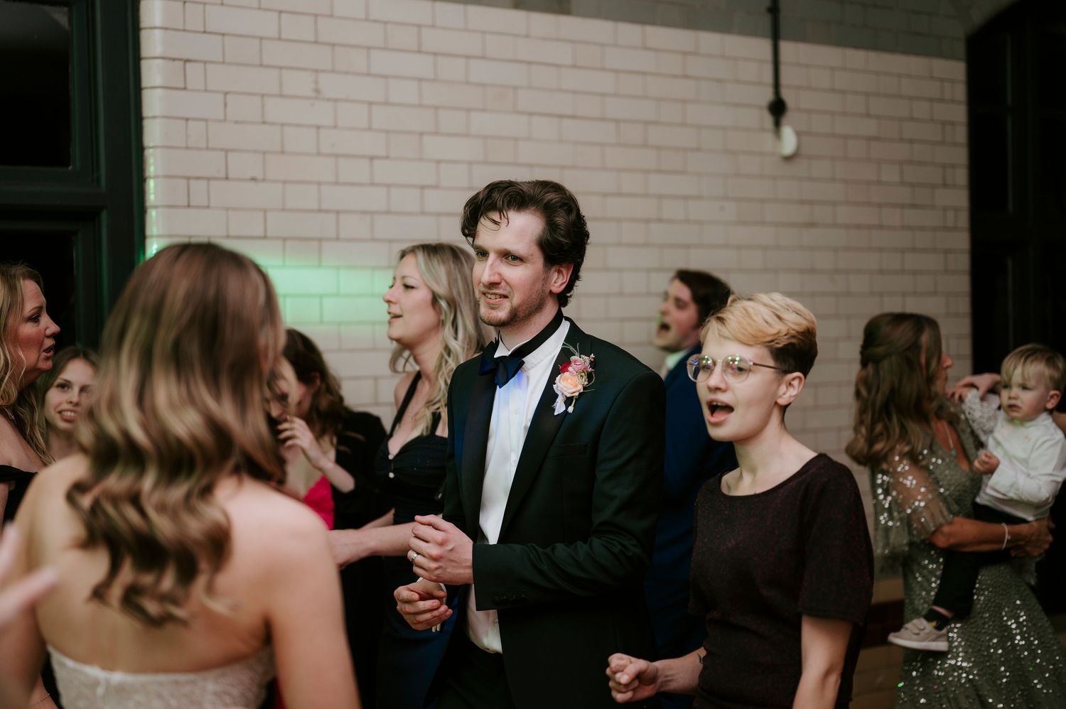 Wedding guests laugh and socialize during an evening reception in a white-walled venue.