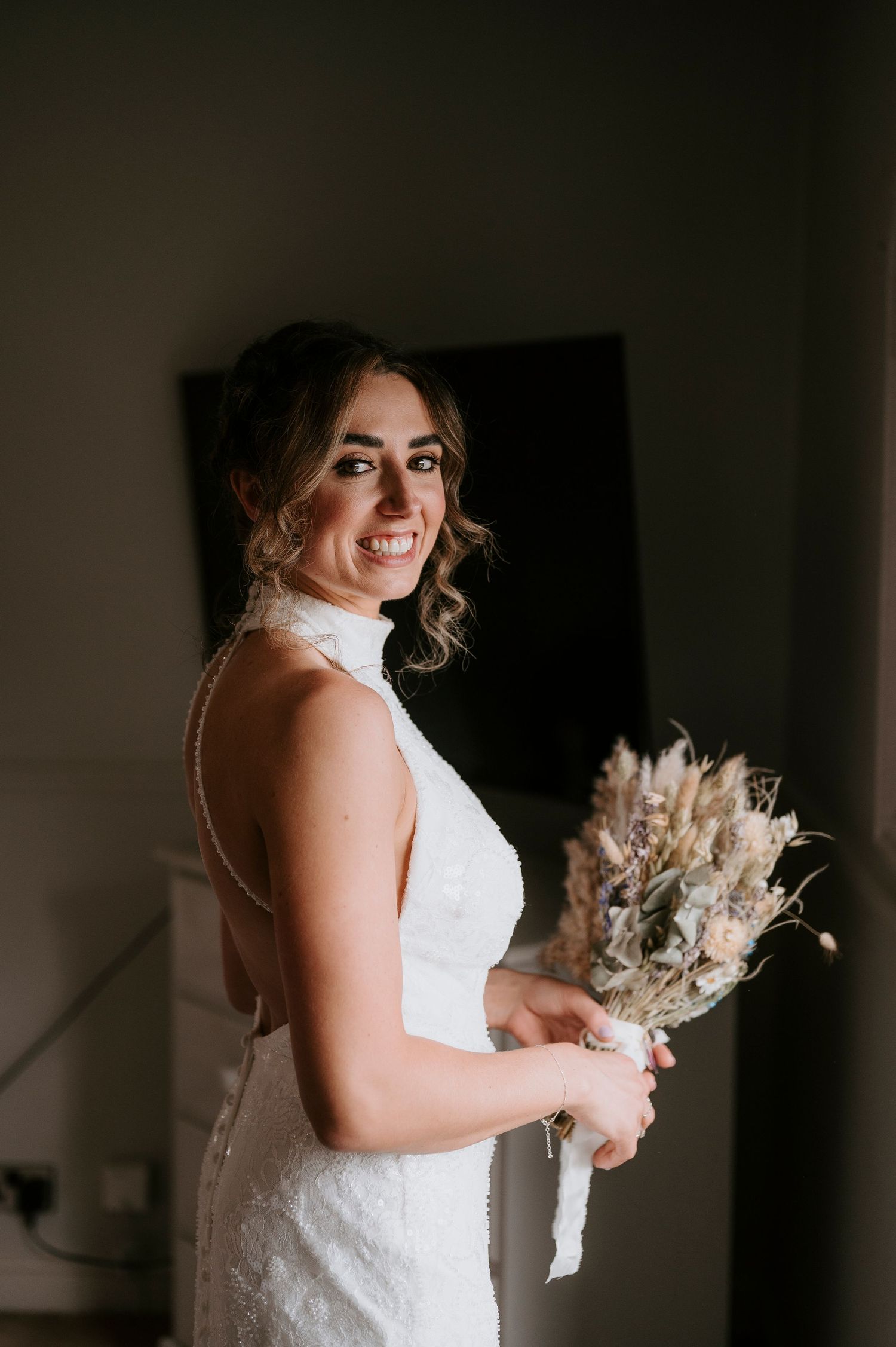A bride in a white lace wedding dress holds a dried floral bouquet in a moody, dark room.