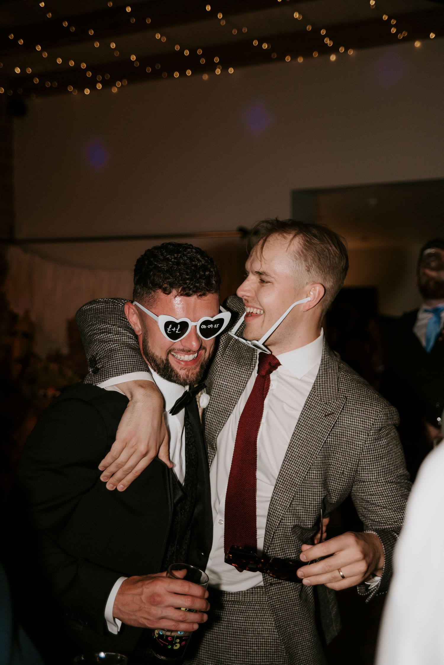Two friends celebrate at a party with one wearing heart-shaped sunglasses and the other in a gray suit with red tie.
