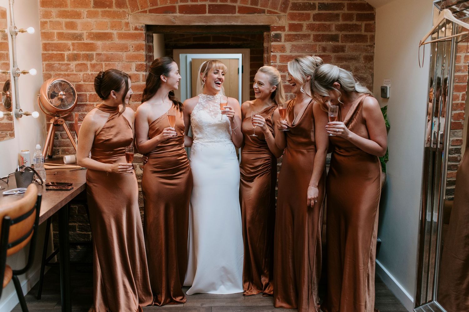 Wedding party in brown bridesmaid dresses laughs together against brick wall at indoor venue.