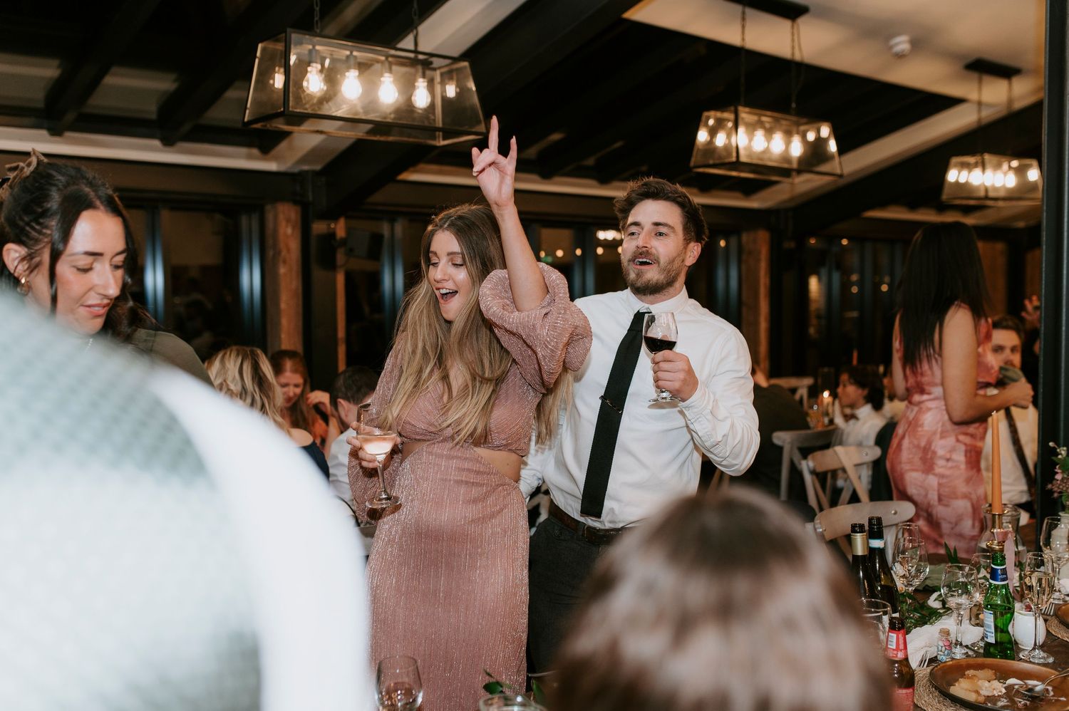 Wedding guests dancing and celebrating at a reception with hanging pendant lights in an indoor venue.
