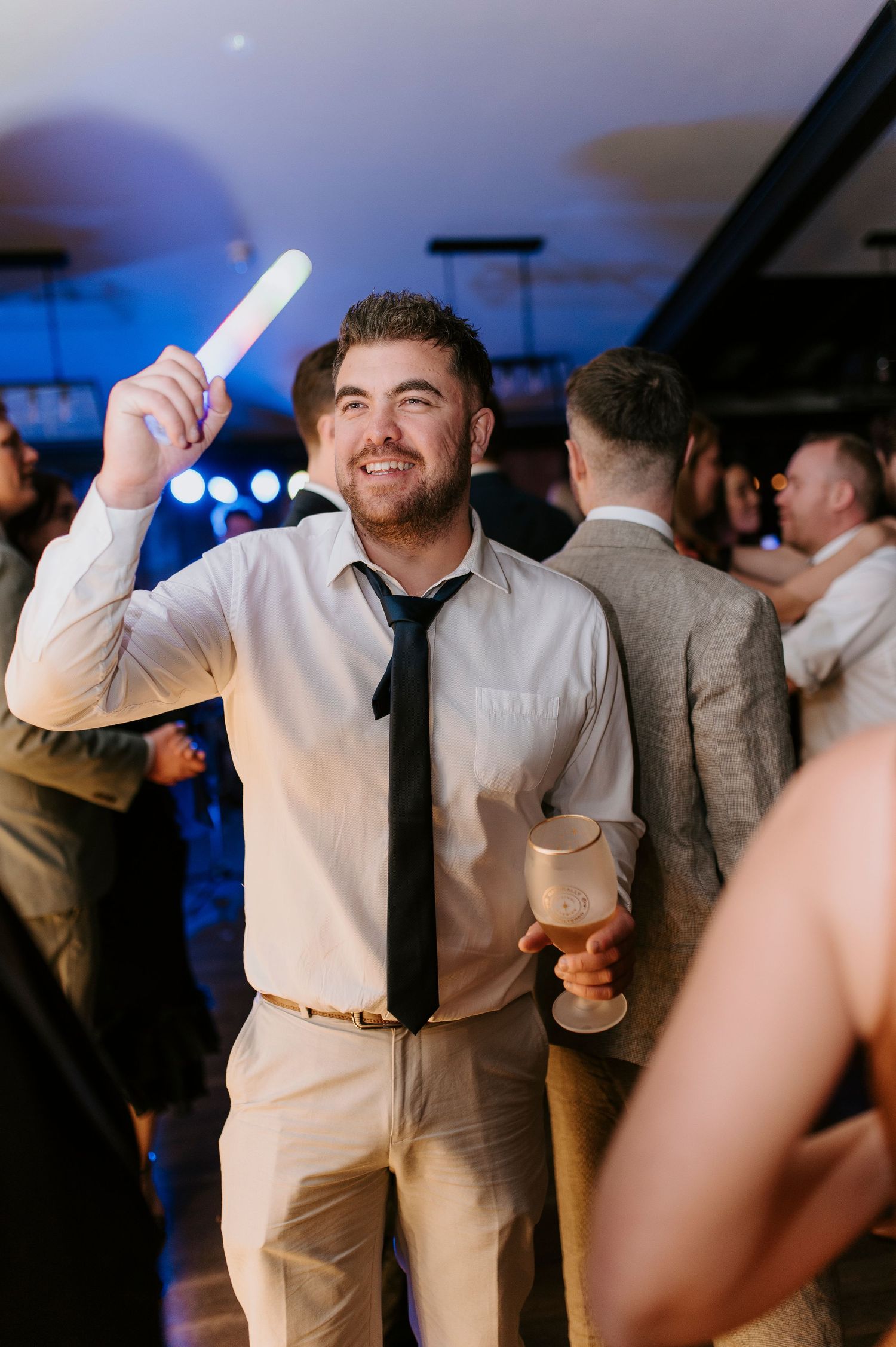 Wedding guests dancing and celebrating at a reception with blue lighting and festive atmosphere.