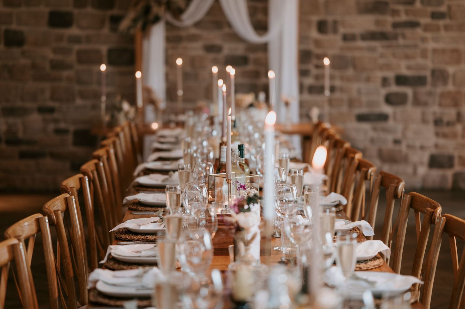 A rustic wedding reception venue with exposed brick walls, wooden chairs, and elegant table settings with glowing candles.