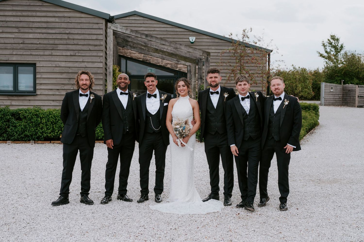 Wedding party in black tuxedos and white gown pose together on a gravel path outside modern wooden buildings.