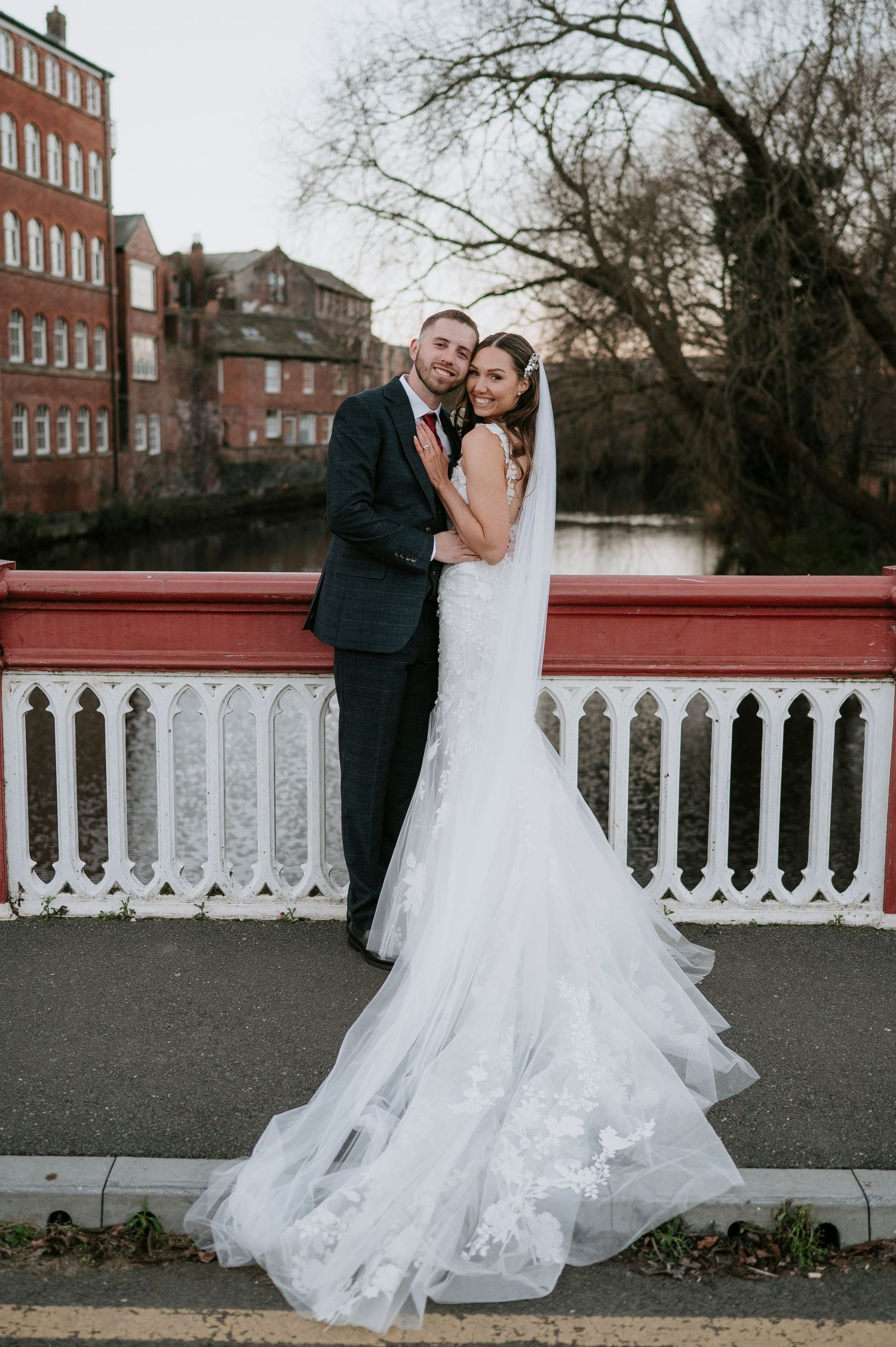 Elegant bride in lace mermaid gown and groom embrace on ornate red and white bridge by river.