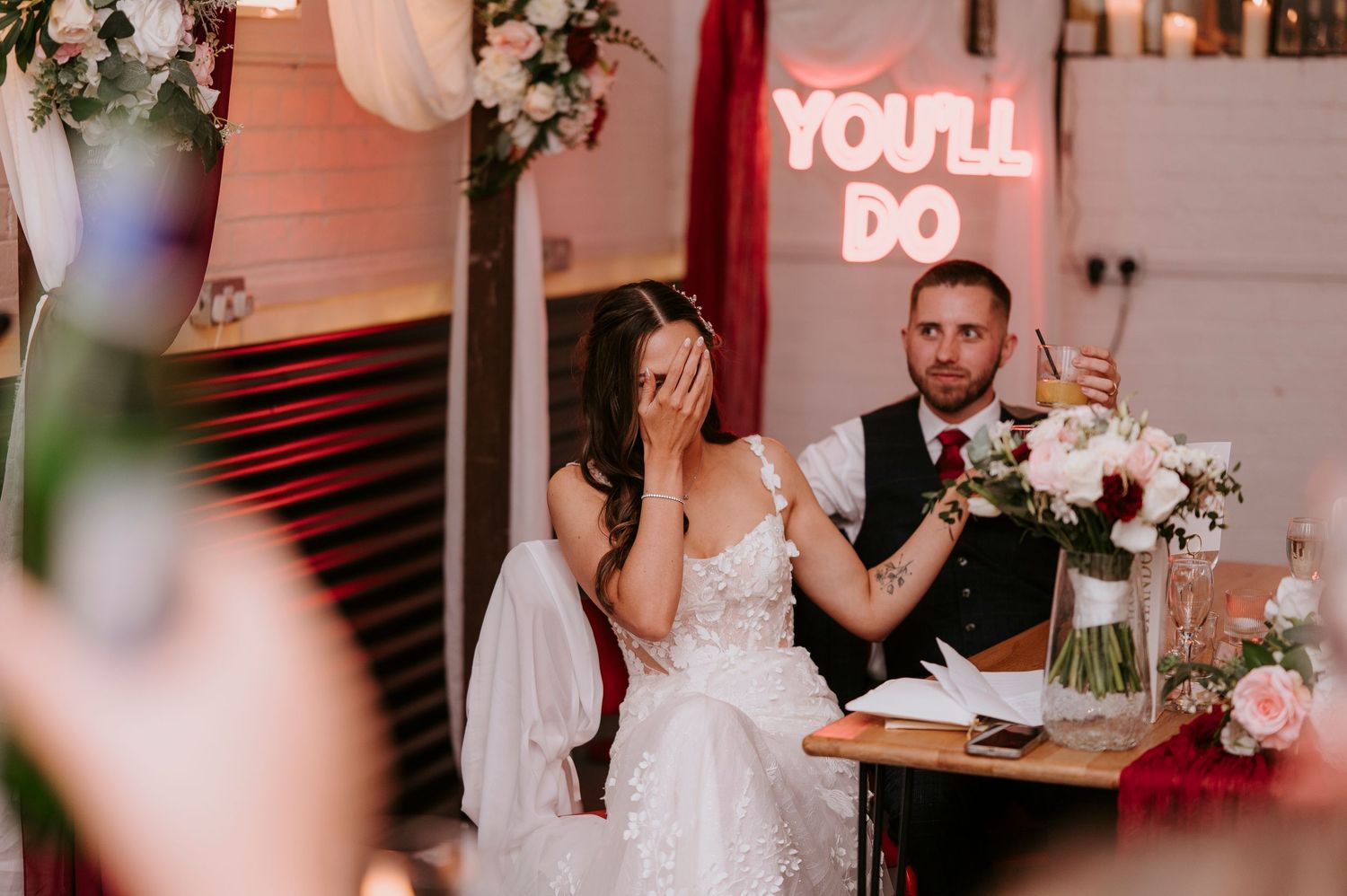 Emotional bride covers face while groom gestures at wedding reception with YOU'LL DO neon sign behind them.