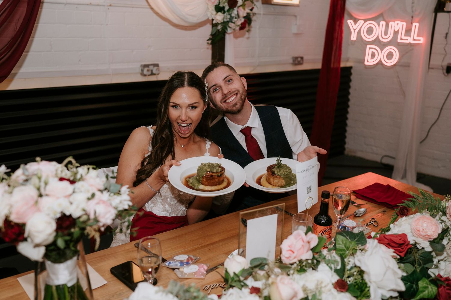 Bride and groom smiling at wedding reception table with floral centerpieces and You'll Do neon sign.