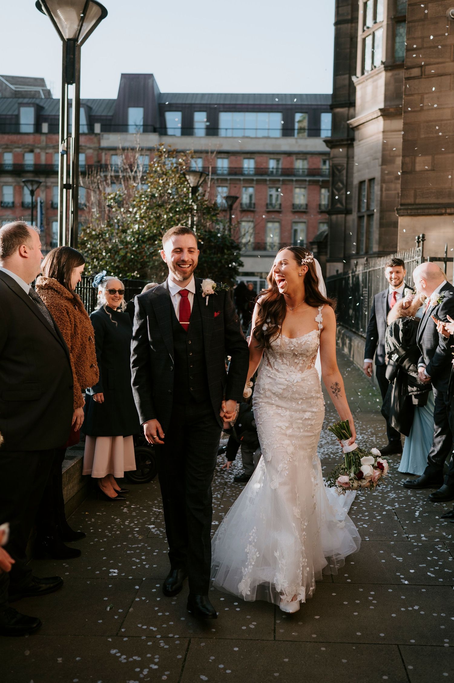 Bride and groom walk through confetti shower outside urban wedding venue, guests cheering around them.