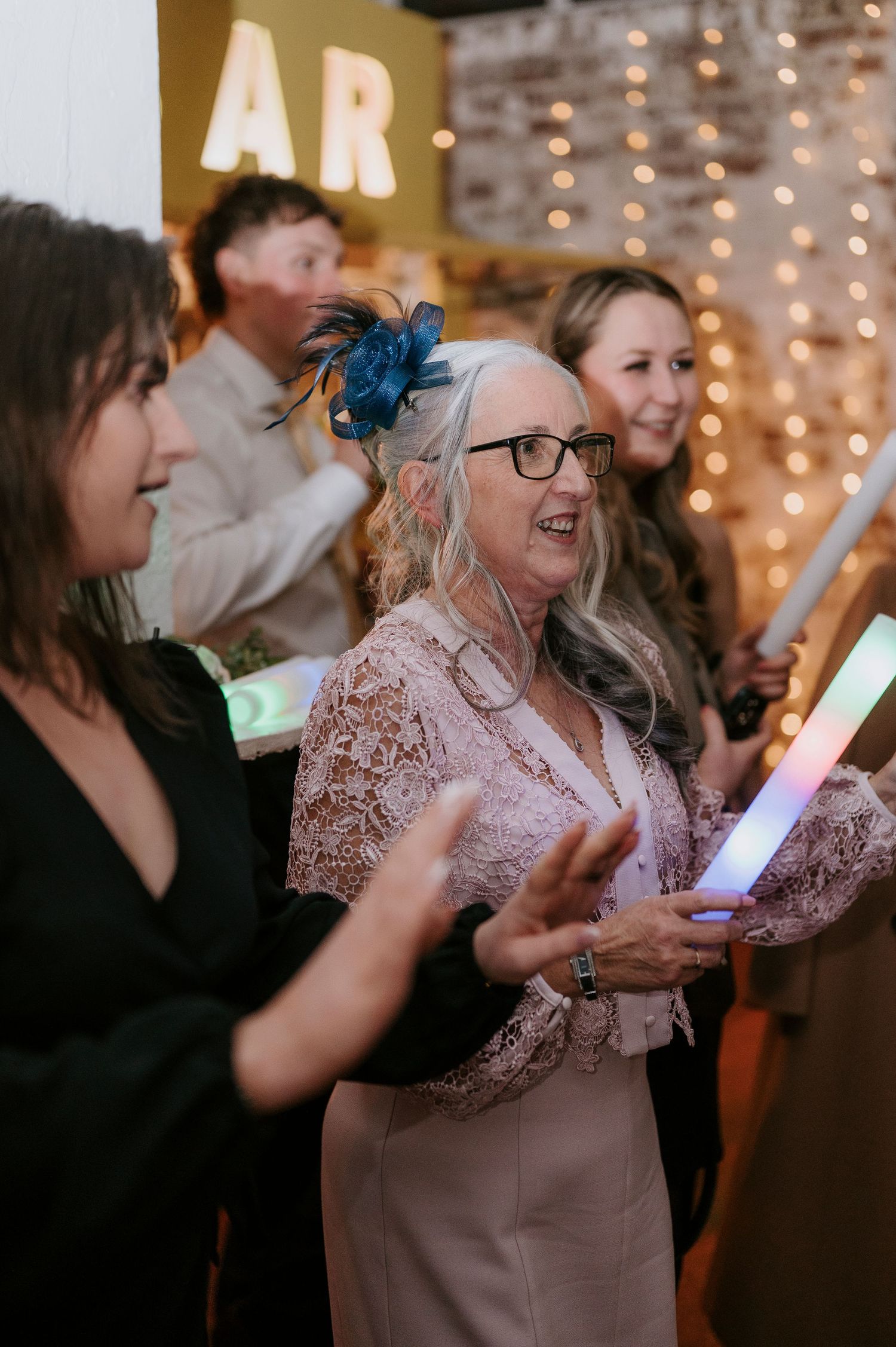 Elderly wedding guest in pink lace dress with blue fascinator dancing with glow stick at reception.