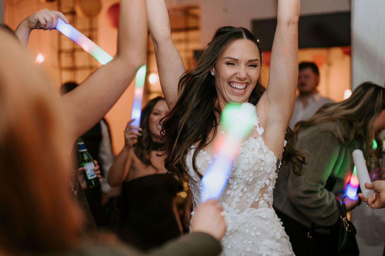 Radiant bride in lace wedding gown dancing joyfully with guests holding glow sticks at reception.