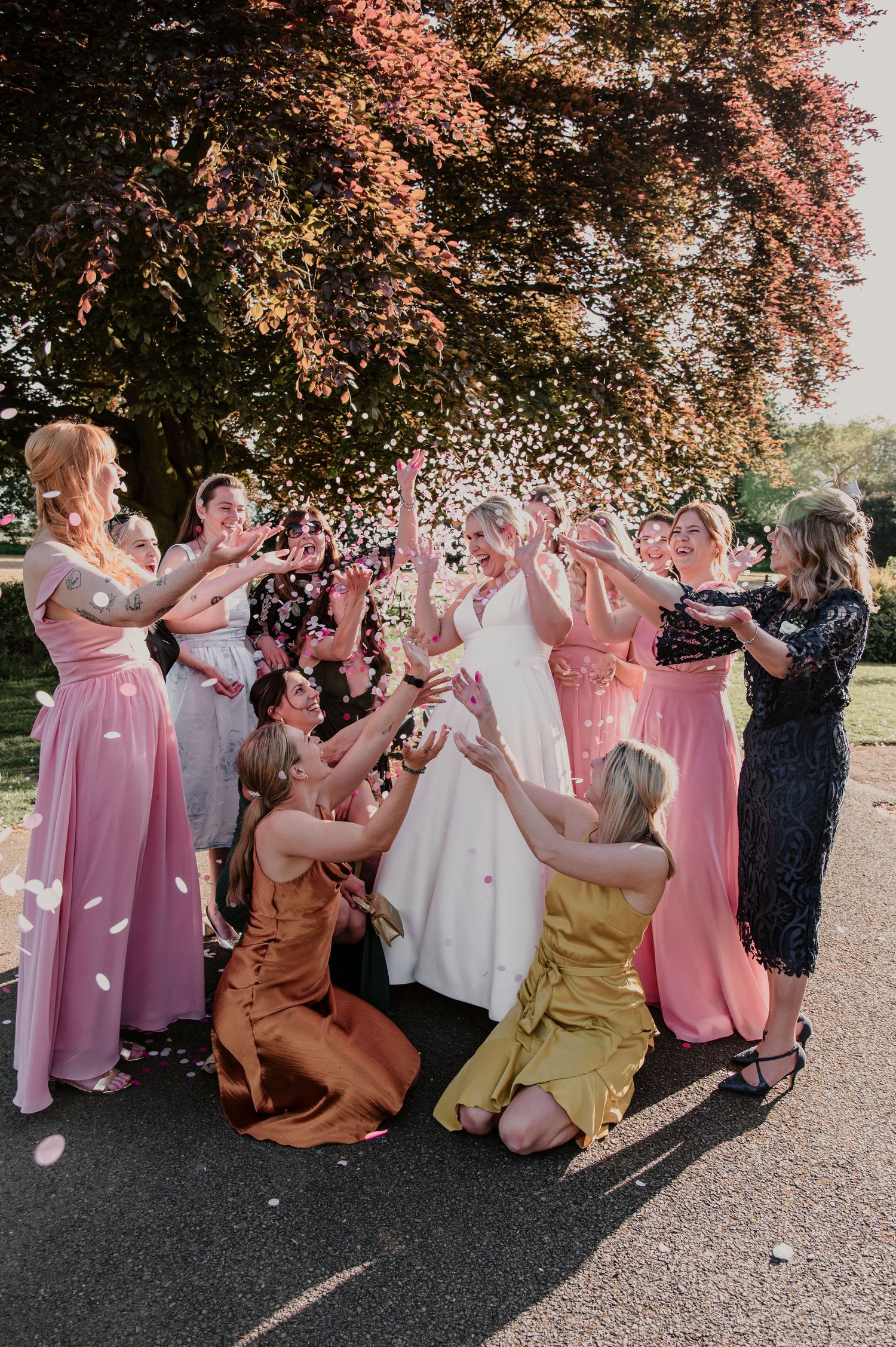Wedding party in pink dresses celebrating and tossing flower petals under a blooming tree.