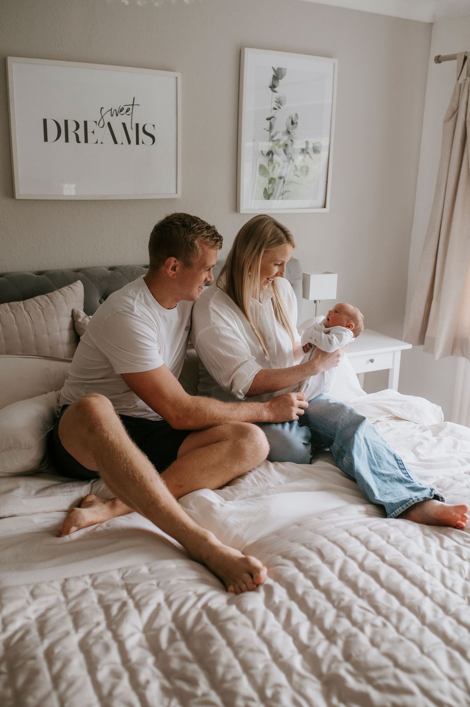 A couple lounges on a white quilted bed with wall art that says 'Dreams' in the background.