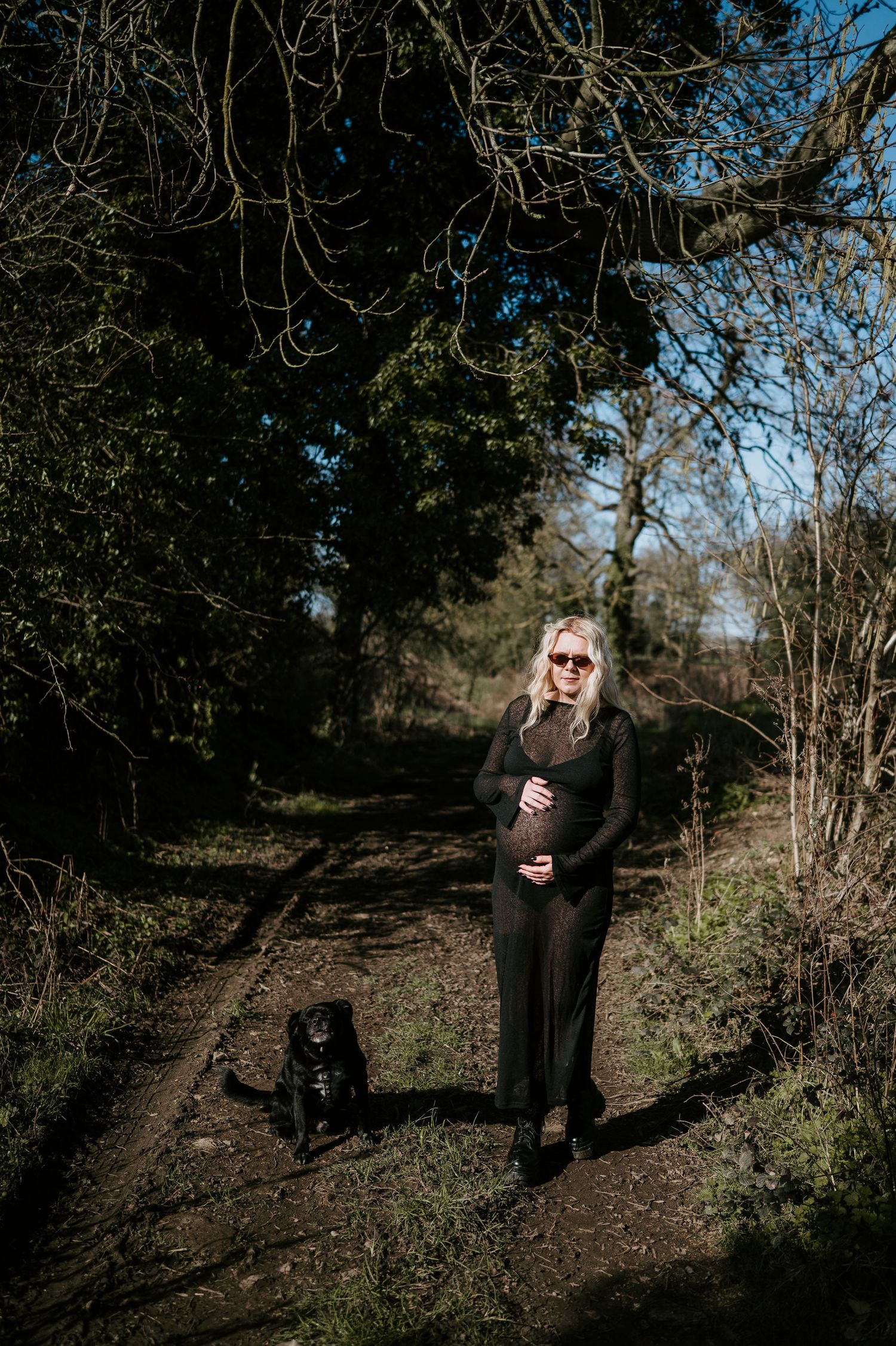 A figure in black stands on a wooded path with a black dog on a sunny day.