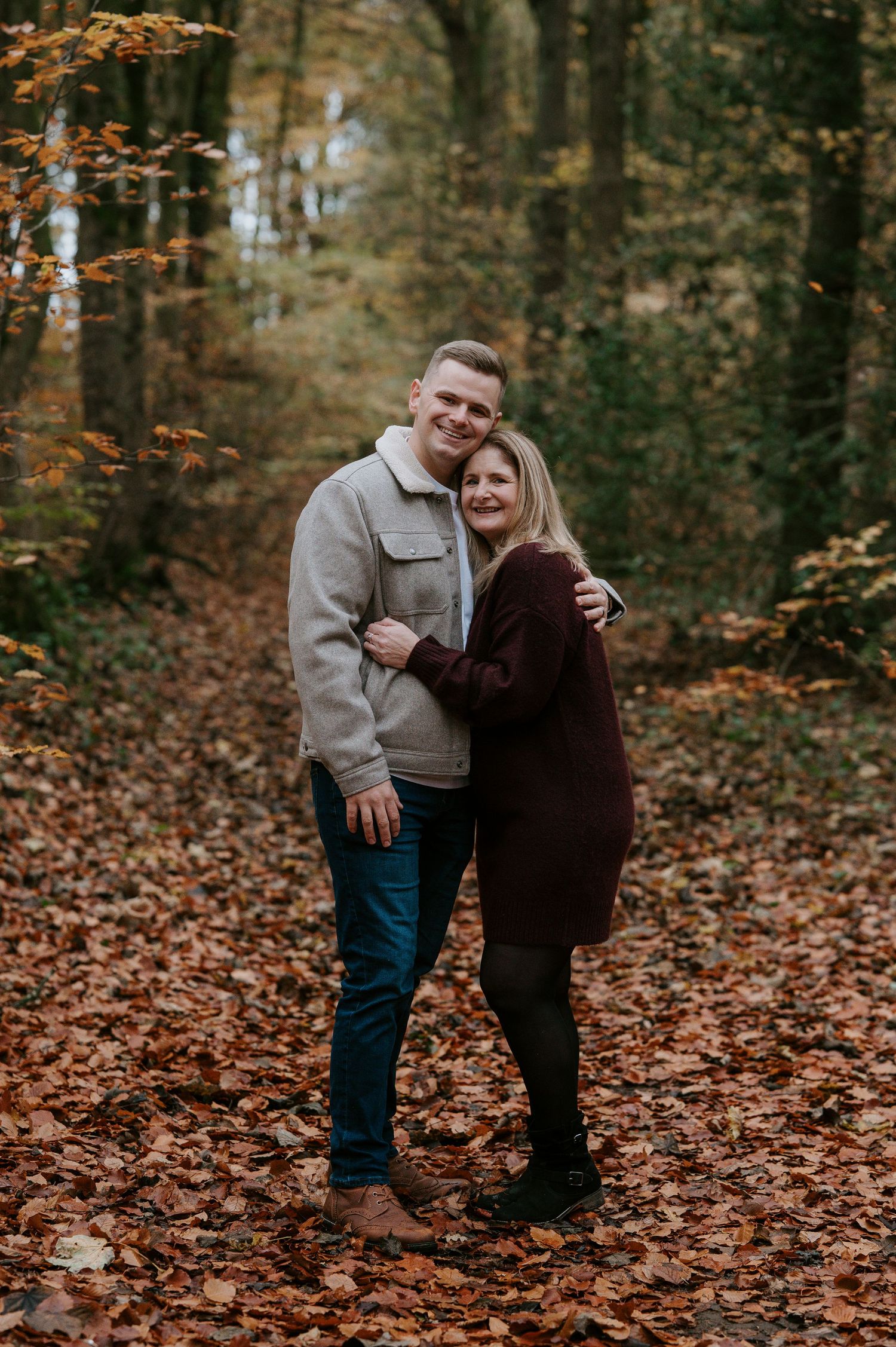 Couple embraces while standing on fallen autumn leaves in a wooded forest setting.