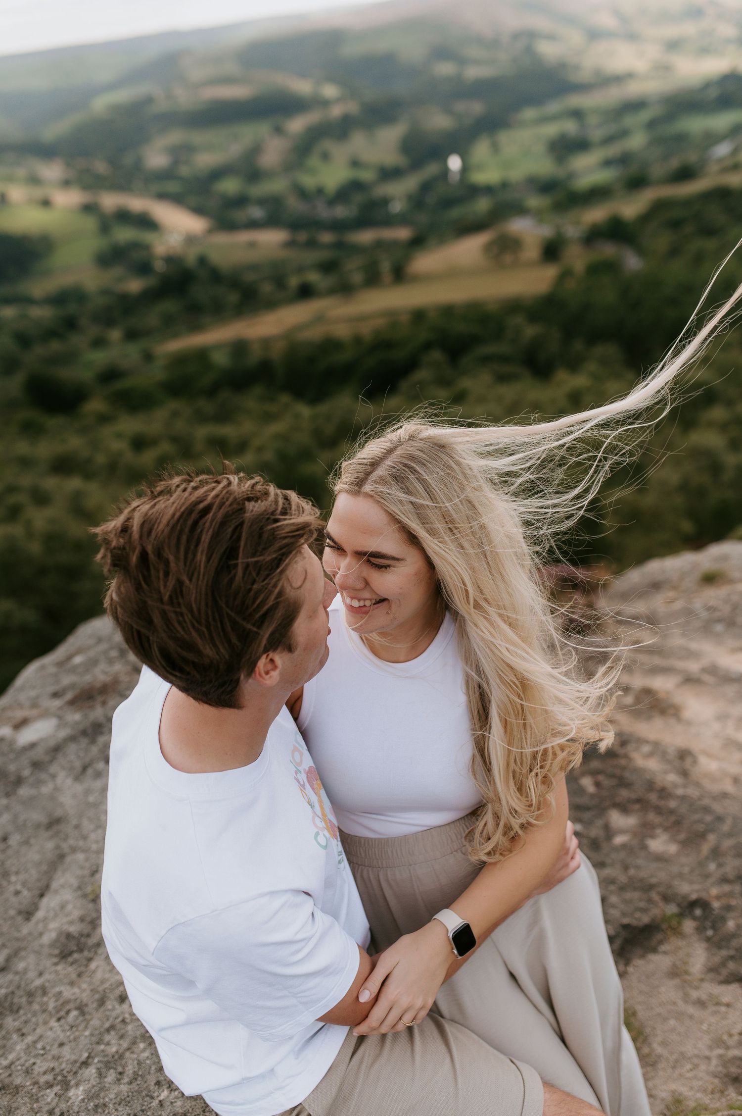 Romantic couple in white outfits sharing intimate moments on a mountain cliff with scenic rolling hills in background.