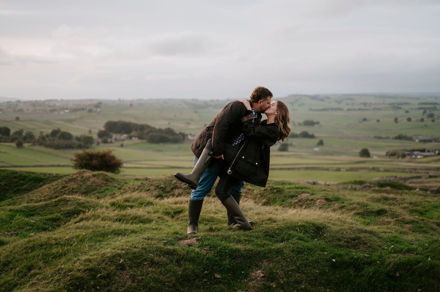 A couple embraces on a hilltop overlooking rolling green countryside on an overcast day.
