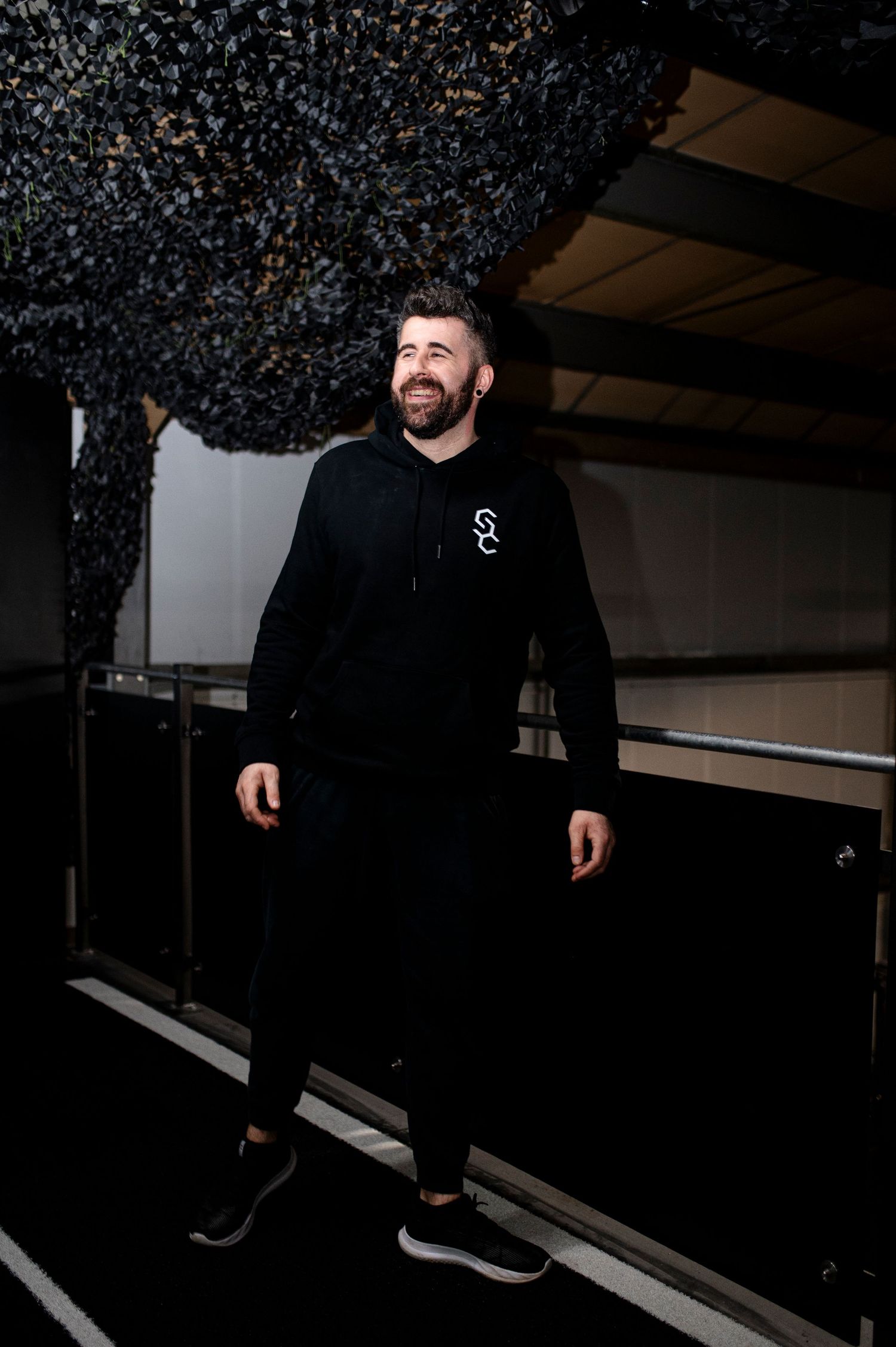 Athletic person in black workout attire smiling while standing in a dimly lit gym setting with textured ceiling.
