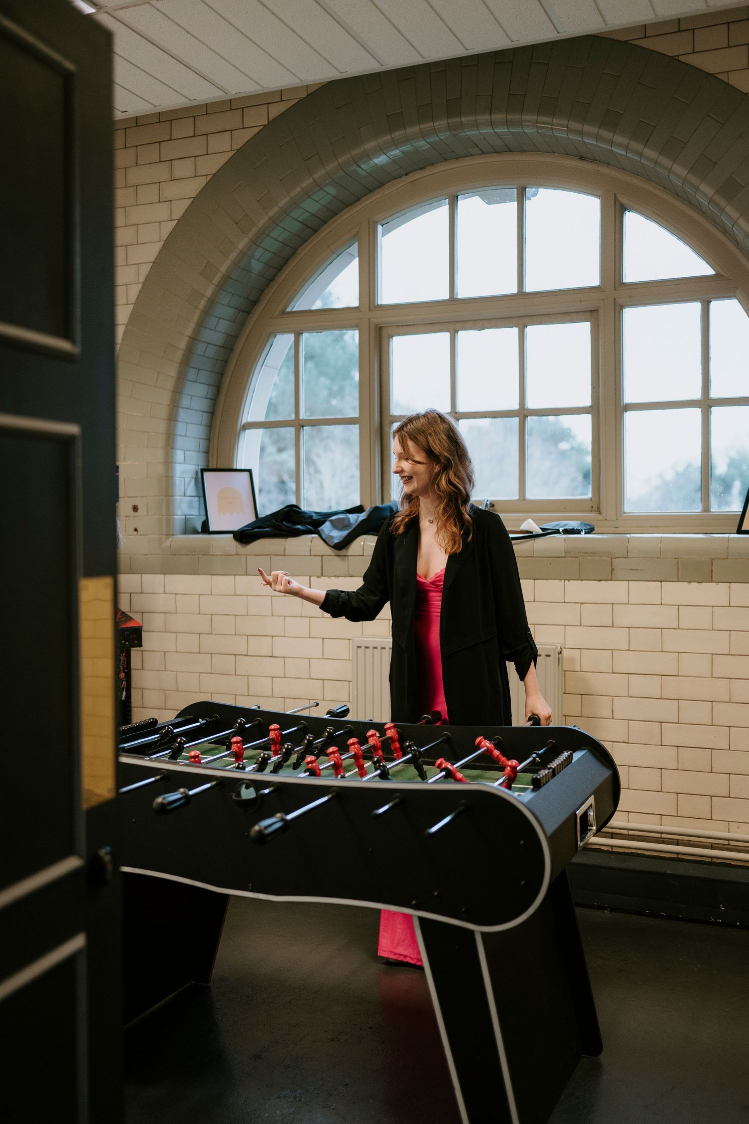 Person in black jacket plays foosball table in room with arched windows and white tiled walls.