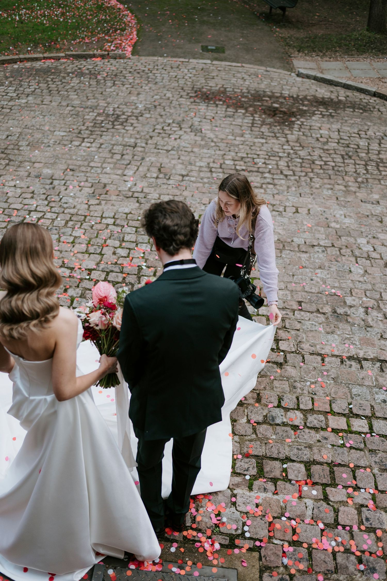 A wedding couple stands on a cobblestone path with pink flower petals scattered around while someone adjusts the dress.