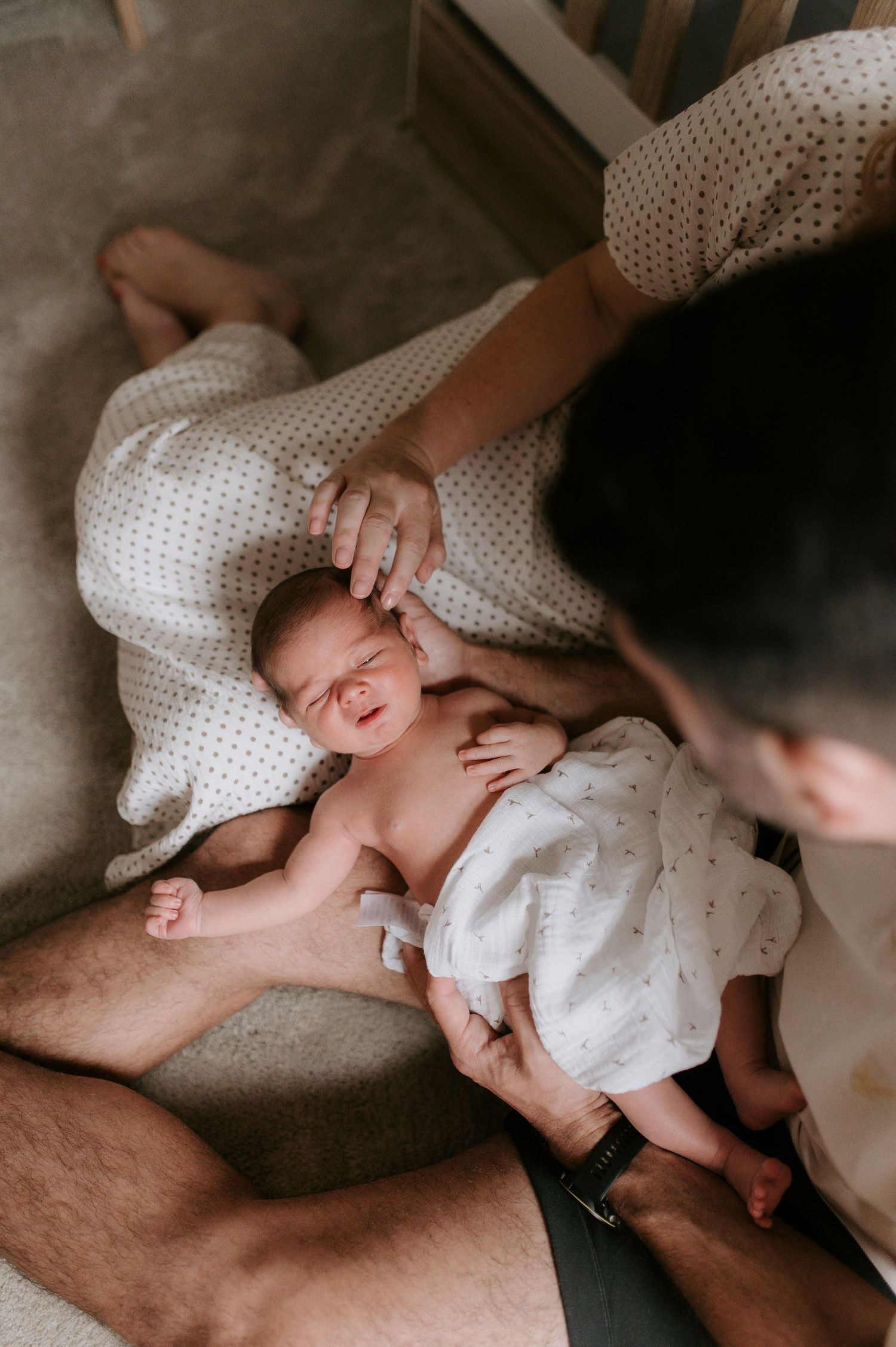 A newborn baby lies on a bed while being gently touched during an intimate family moment.