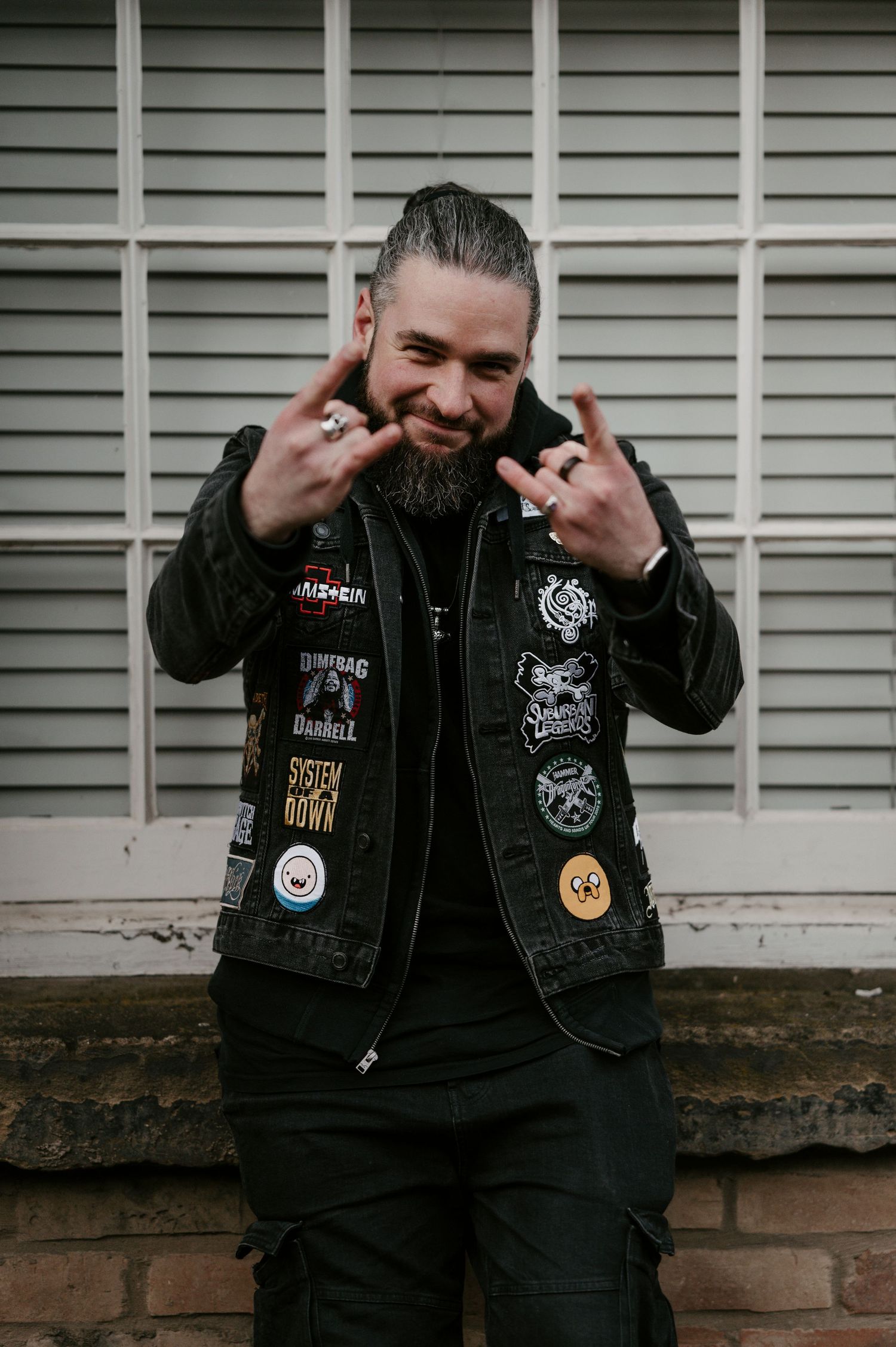 Person wearing black jacket with patches making rock gestures against slatted wall.