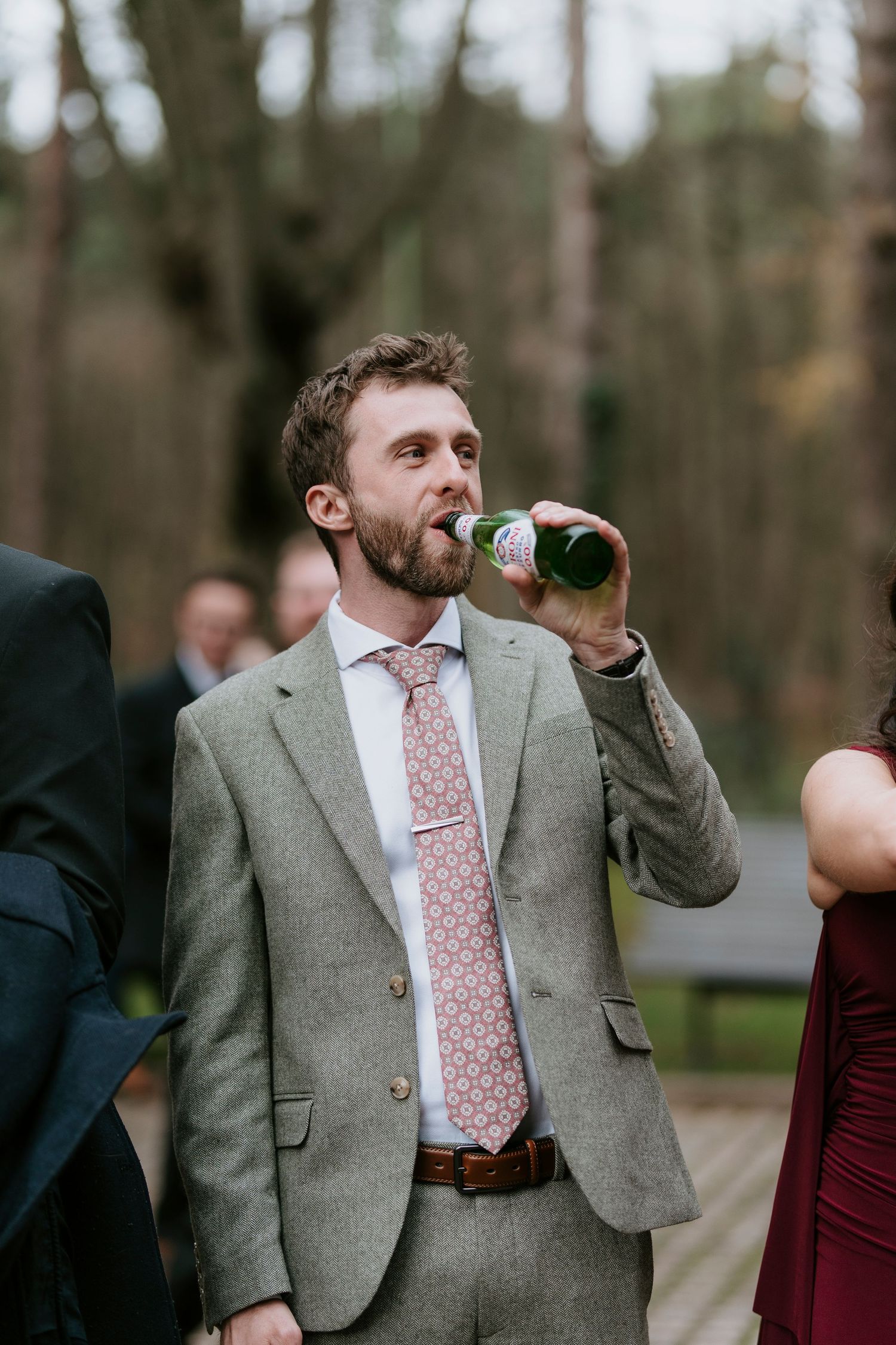 A person in a gray suit and pink tie drinks from a bottle at an outdoor event.