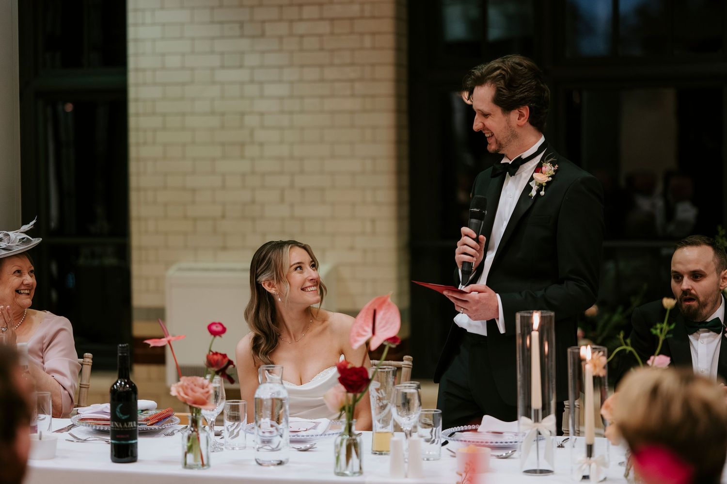 Wedding reception guests seated at long tables with white linens and floral centerpieces in rustic brick venue.