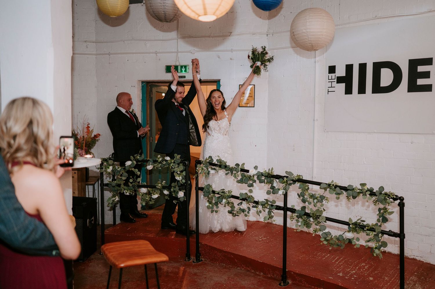 Joyful newlyweds make grand entrance through doorway at industrial wedding venue decorated with colorful paper lanterns.