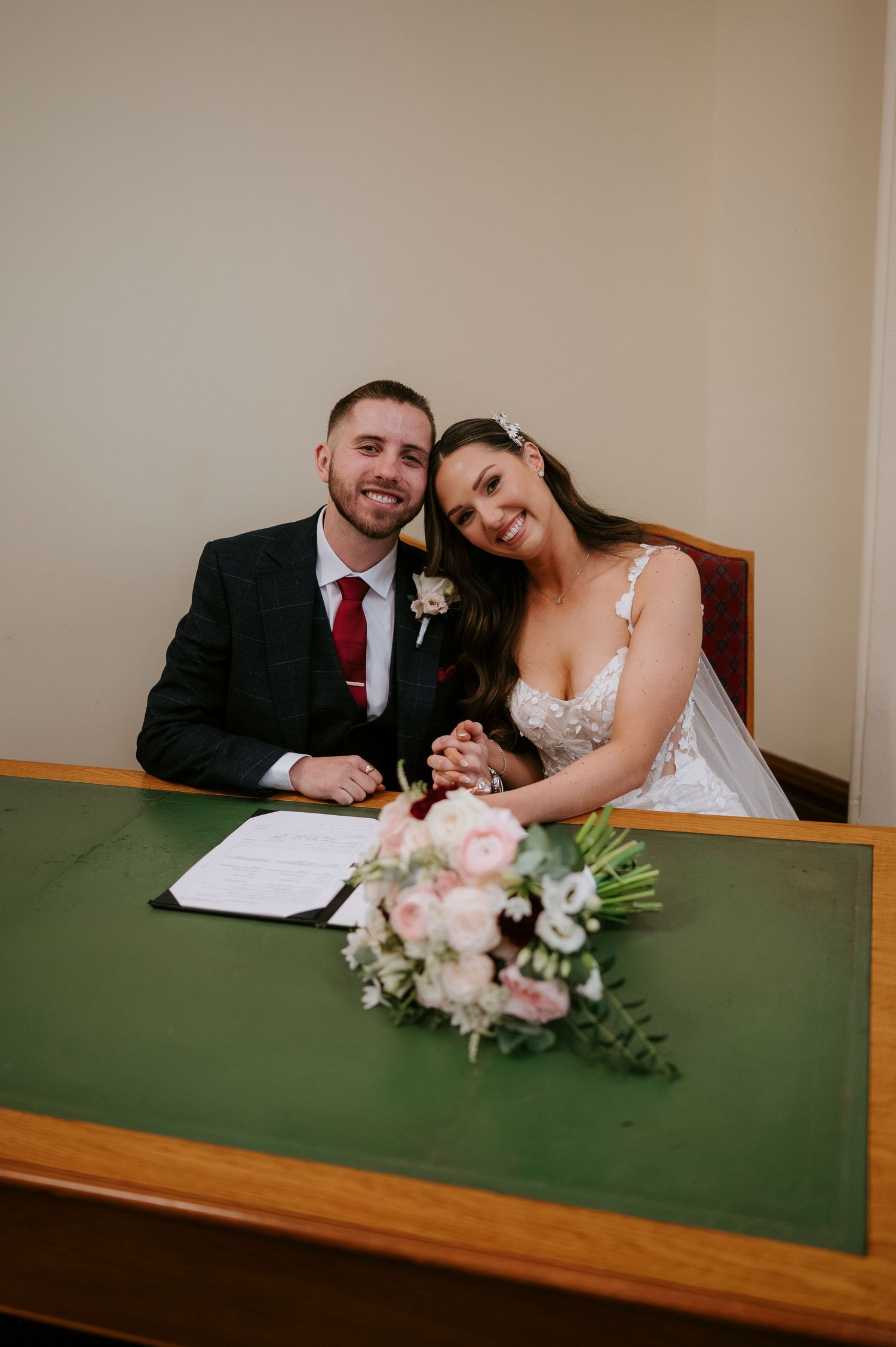 Newlywed couple smiling at signing table with wedding bouquet of pink and white flowers during marriage ceremony.