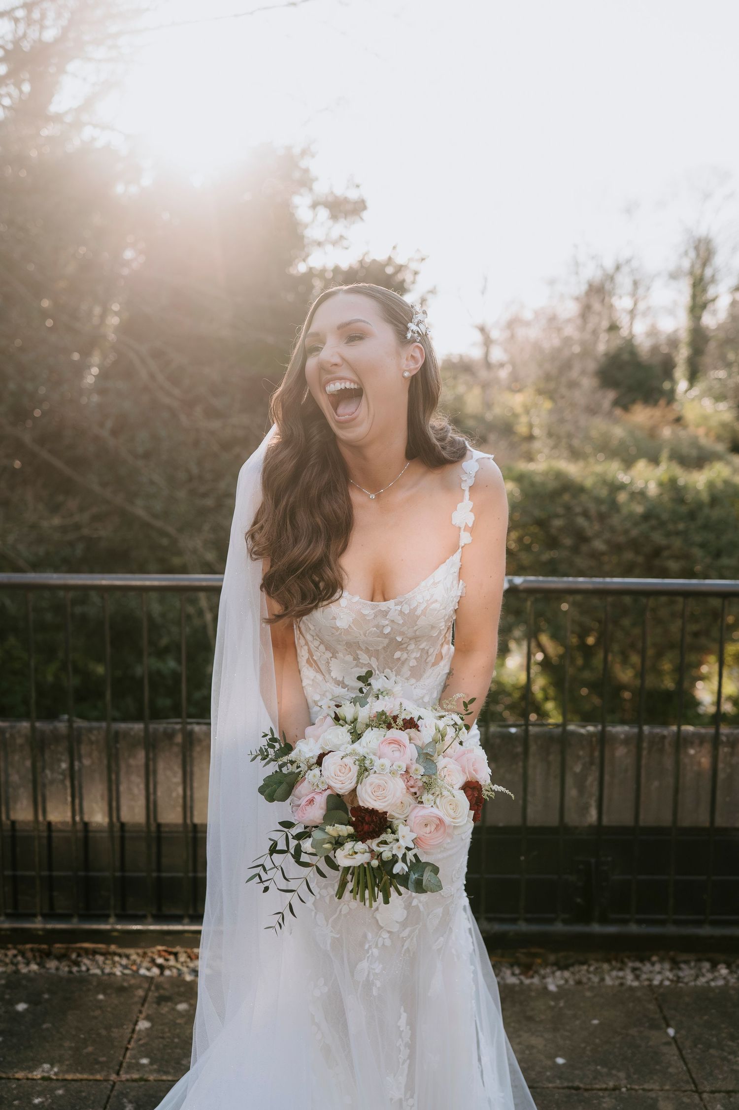 Joyful bride in lace gown holding pink and white bouquet, laughing outdoors in golden sunlight with veil and floral hair piece.