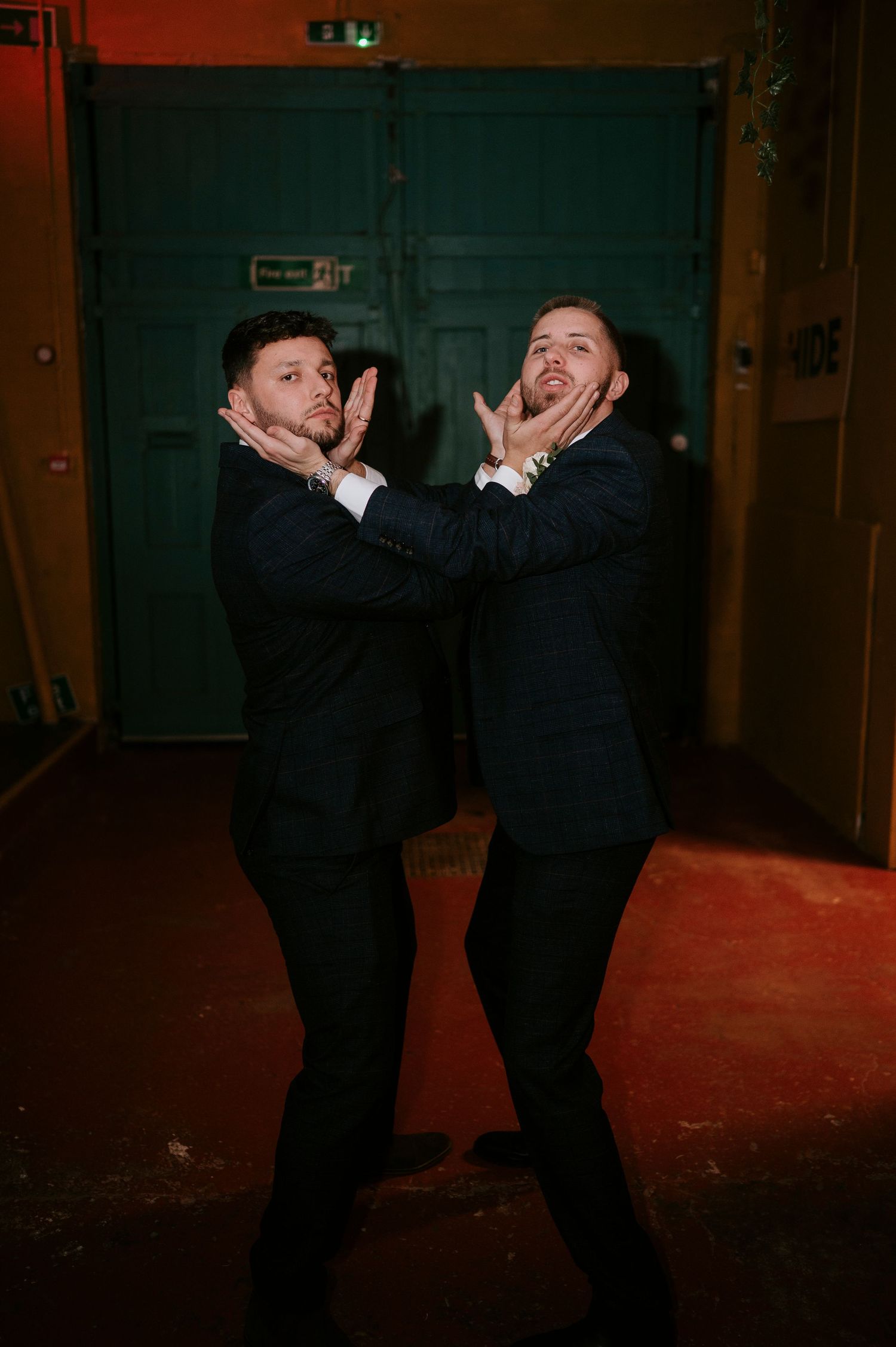 Two men in dark suits playfully posing together in front of teal double doors at a nighttime event.