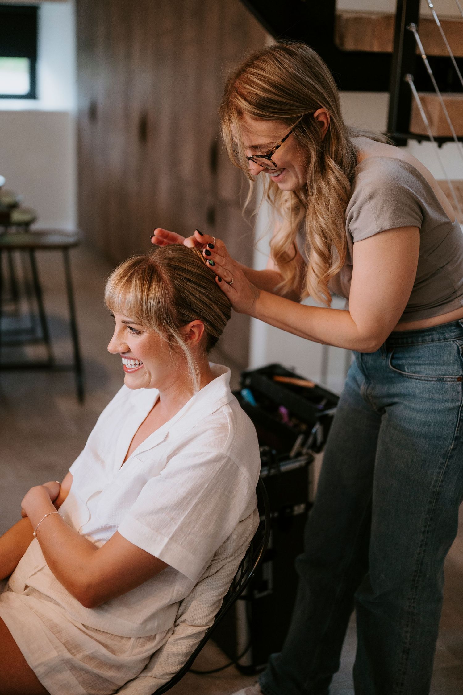A hairstylist in jeans and glasses styles a client's hair in a white robe during wedding preparations.