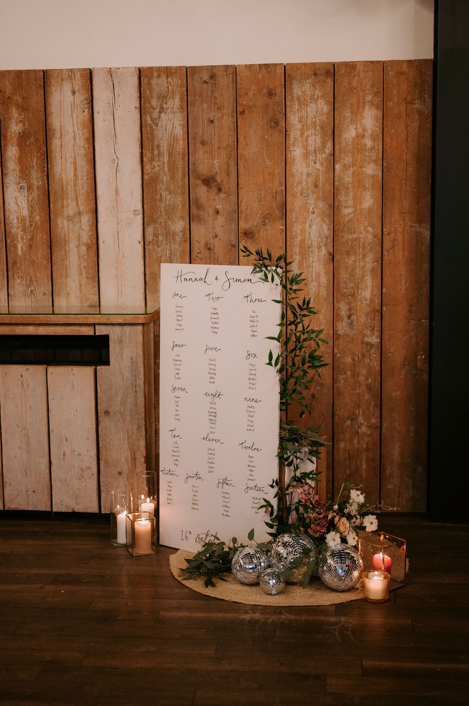 Wedding seating chart display with white board, candles and floral decorations against rustic wooden wall backdrop.