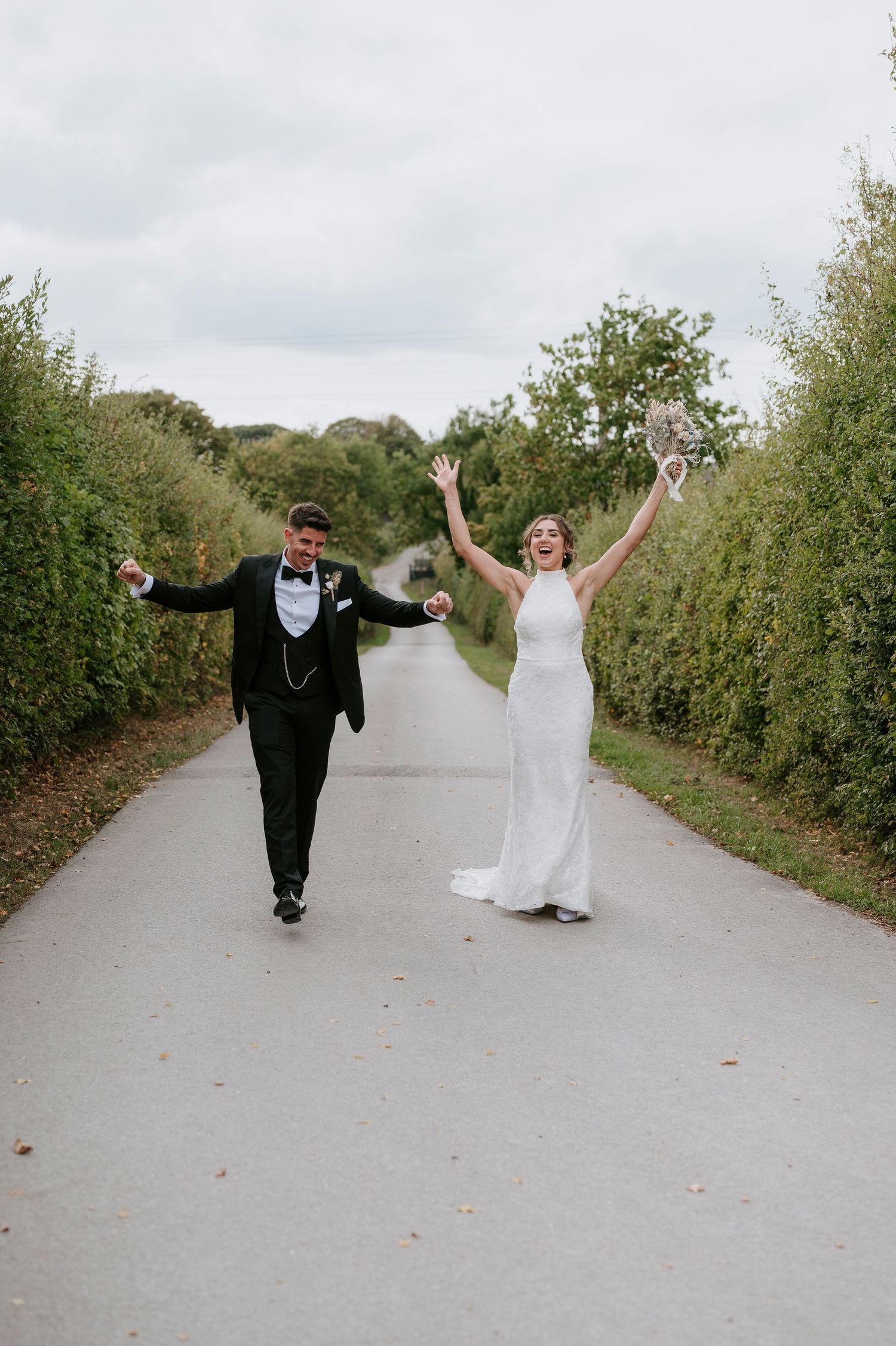 A couple in formal wedding attire playfully runs and dances together on a country path lined with green hedges.