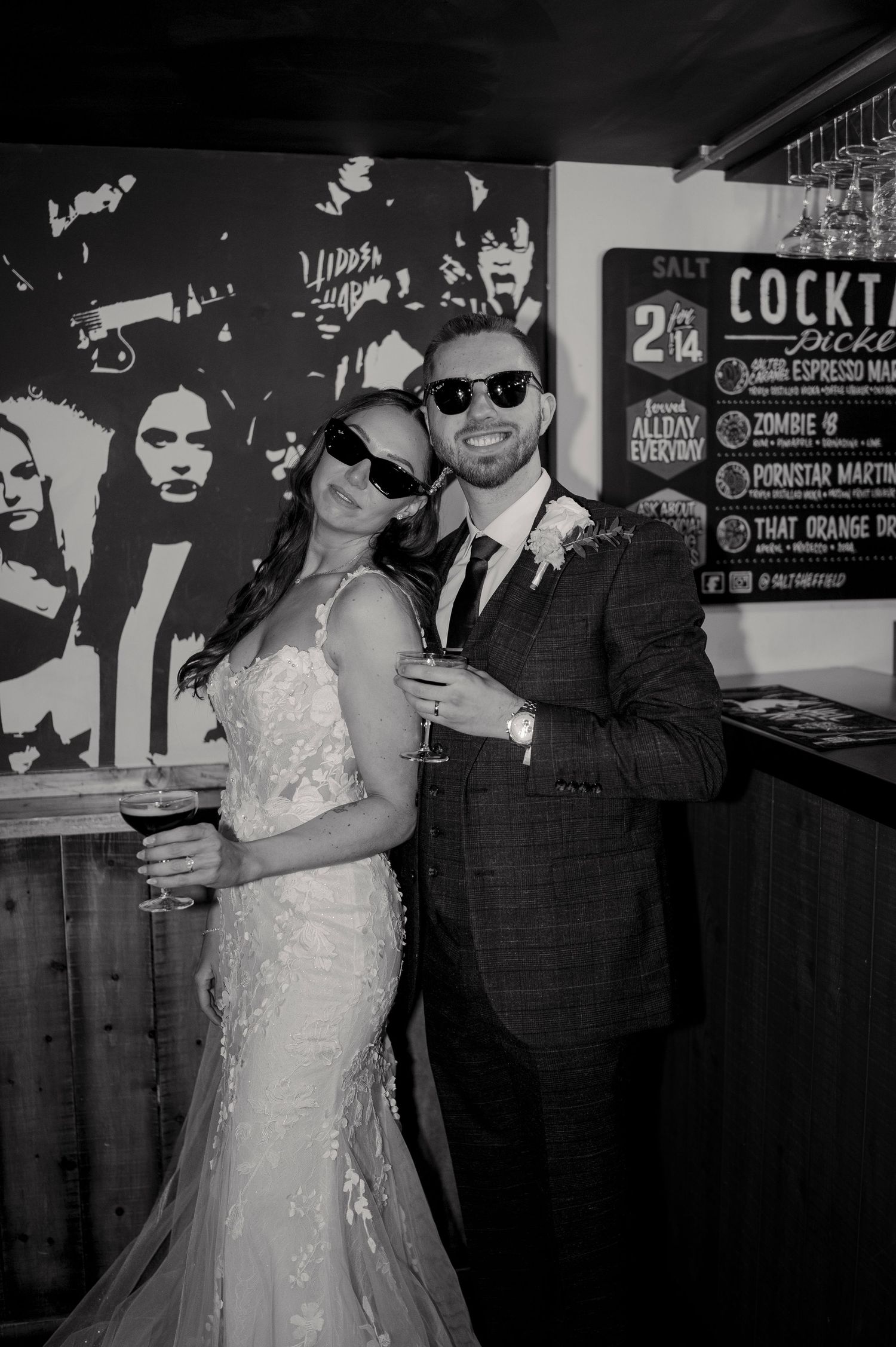 Bride and groom pose playfully in sunglasses at a bar in black and white wedding photo.