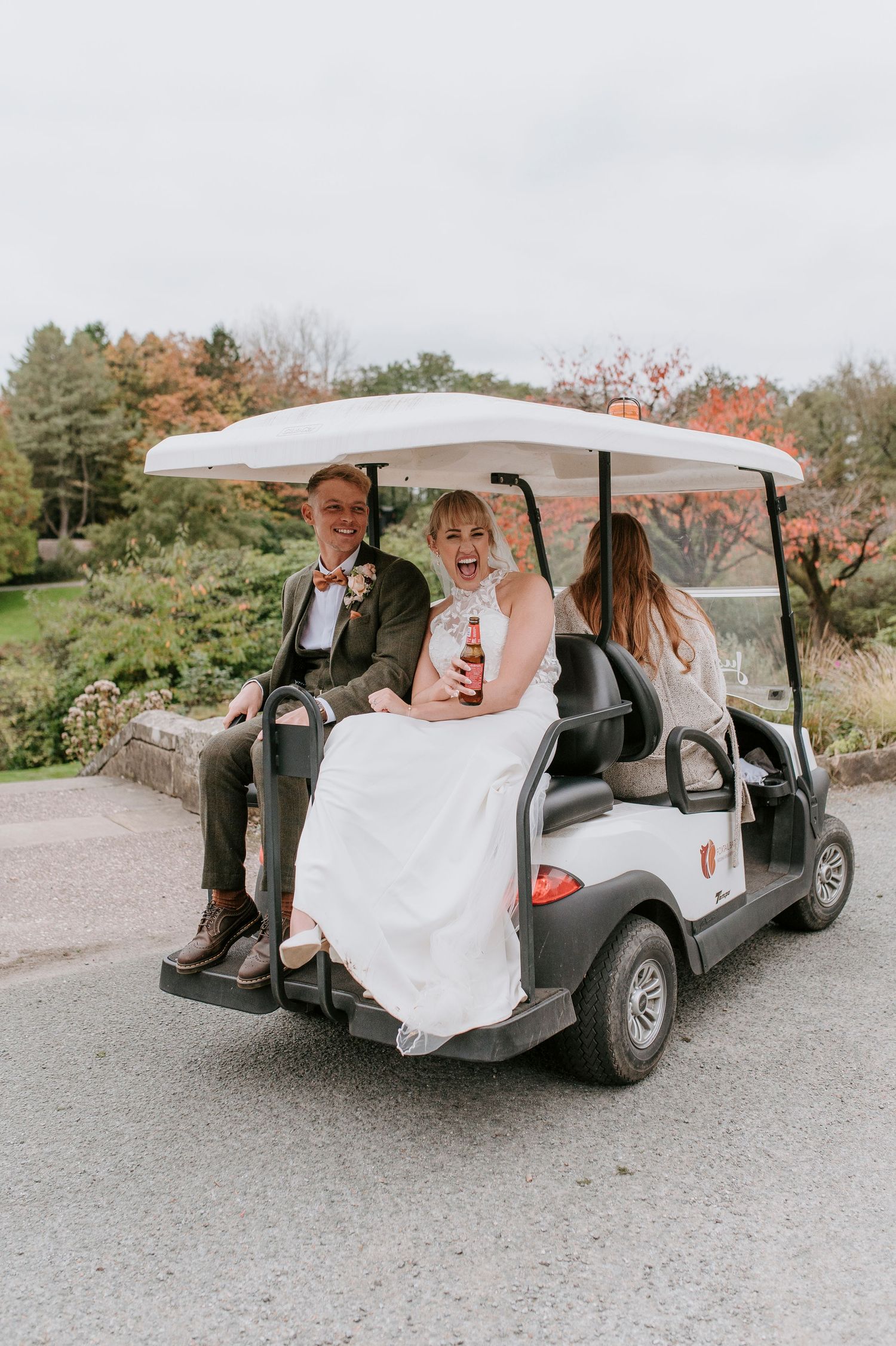 Wedding party rides in a white golf cart along a path surrounded by autumn trees and greenery.