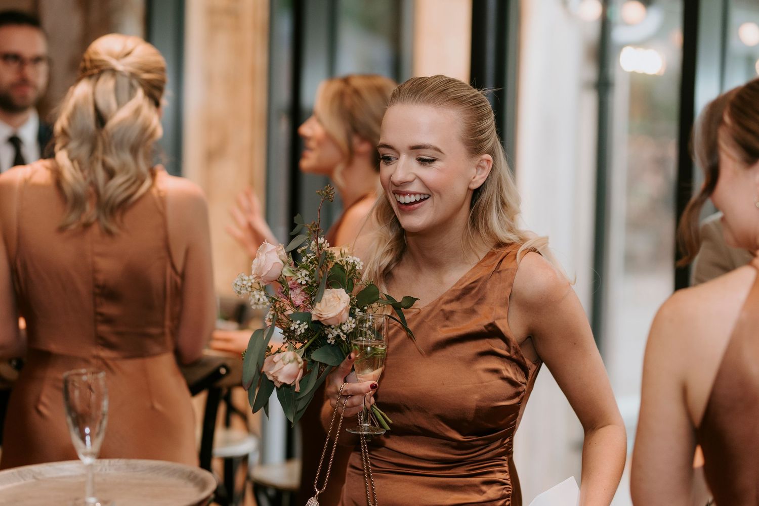 Bridesmaids in matching brown satin dresses laugh together during a wedding celebration.