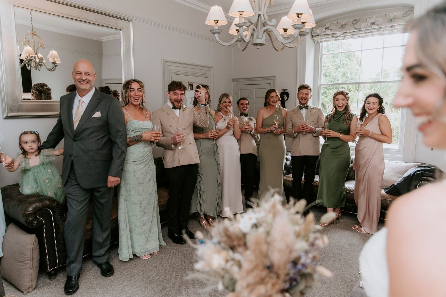A wedding party in pastel dresses stands in line against a wall beneath elegant chandeliers in a bright indoor setting.