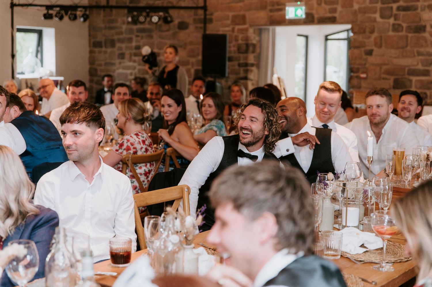 Wedding guests seated at long tables in a rustic brick-walled venue enjoy dinner and conversation during reception.